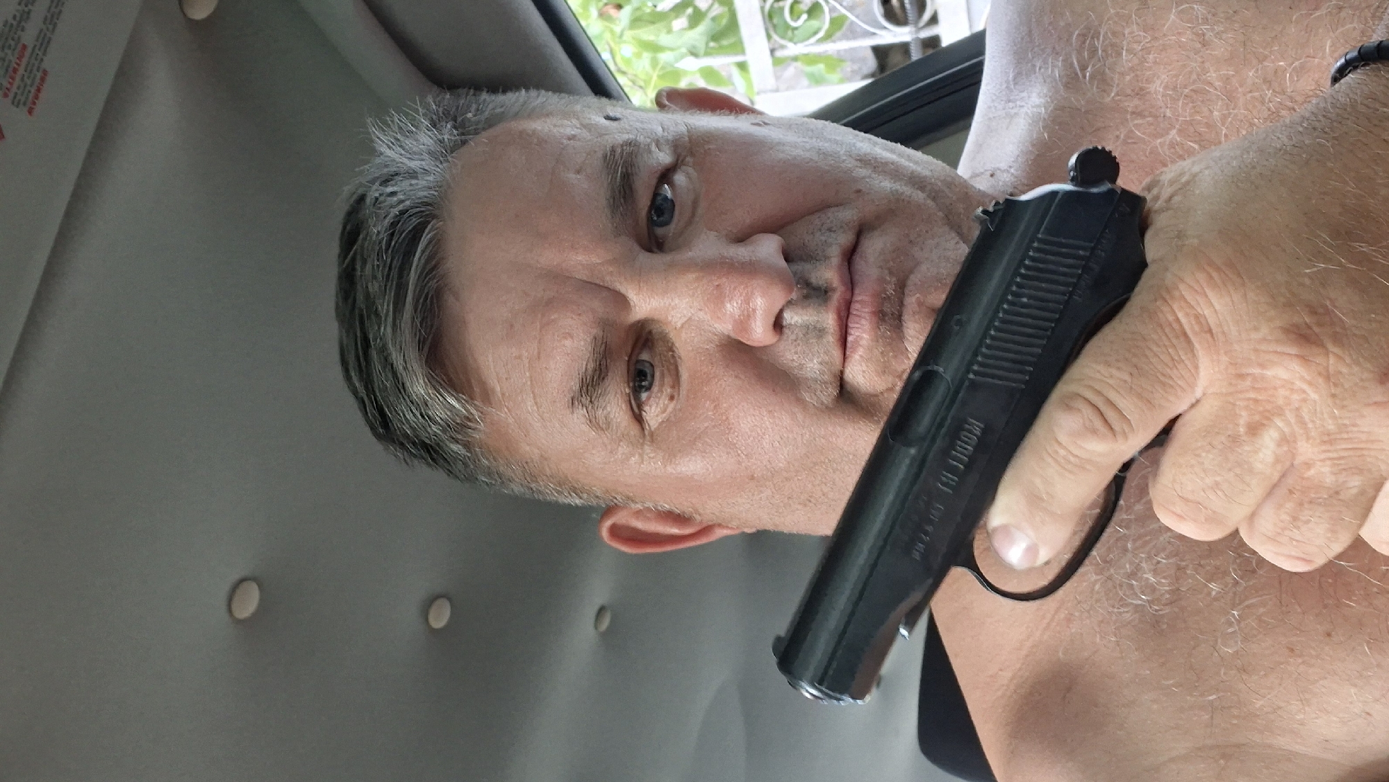 A shirtless, middle-aged man with short, graying hair is sitting in a vehicle. He is holding a black semi-automatic pistol in his right hand, pointing it towards the camera. The man's skin is fair, and he has a few moles visible on his face and chest. The interior of the vehicle is dimly lit, with the ceiling visible in the background. Outside the vehicle, green foliage and a white fence can be seen. The man's facial expression is serious. The image is a close-up, focusing on the man's upper body and the weapon he is holding. The location in the city of Voinova, Moldova, is not depicted in the image.