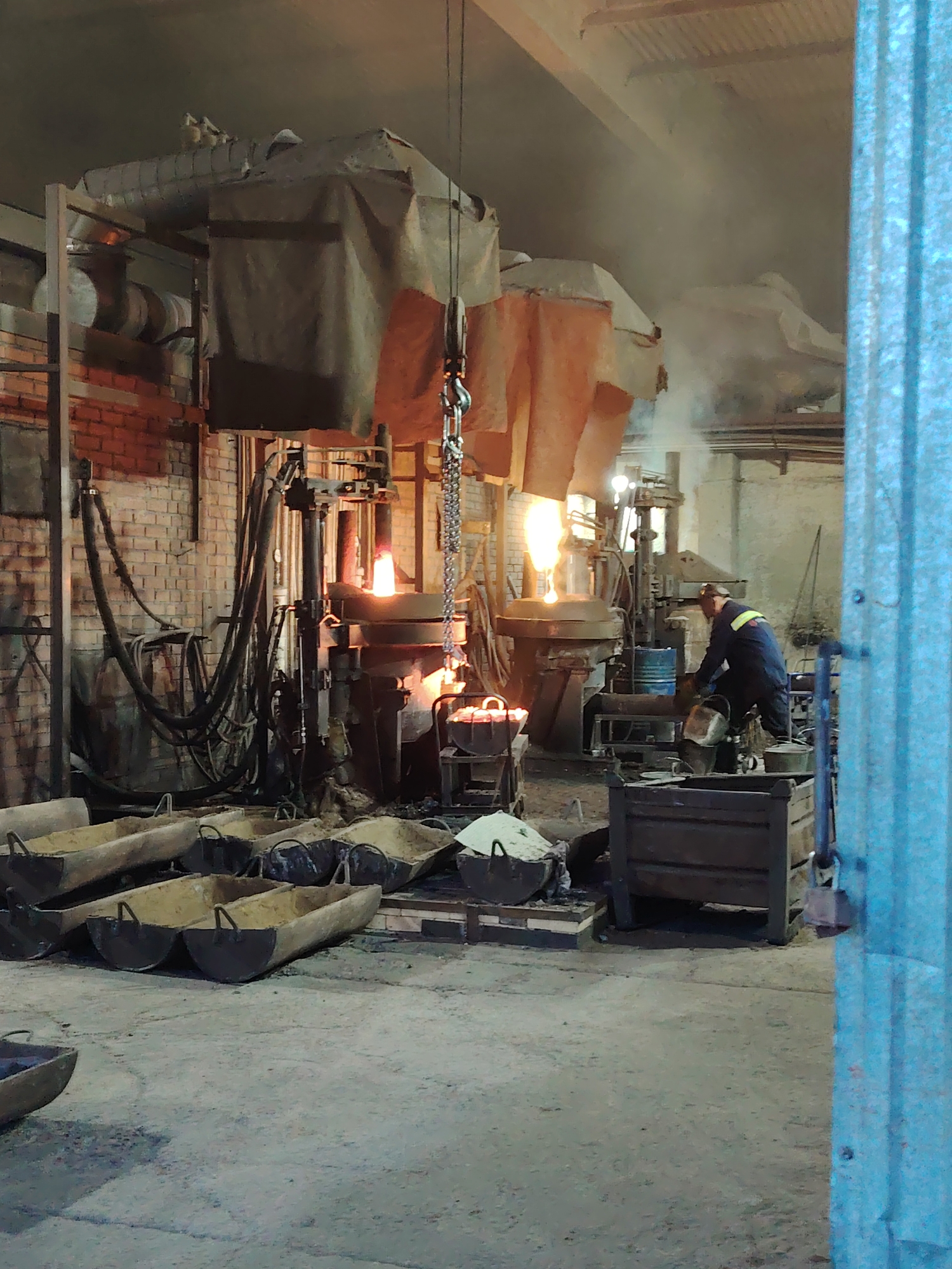 The image shows a metal foundry in Noviy, Russia. Two industrial furnaces are visible, emitting molten metal and smoke. A worker in dark overalls and a yellow safety stripe is tending to one of the furnaces. In the foreground, several metal troughs filled with sand are arranged on the floor. A brick wall is visible in the background, along with ventilation equipment. The light is dim, creating a stark contrast between the glowing molten metal and the surrounding environment.