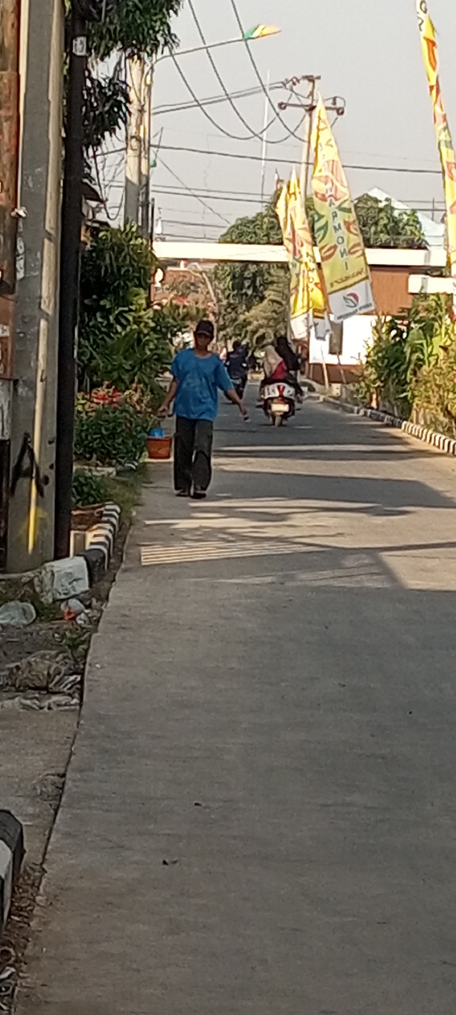 In the city of Lubangbuaya, Indonesia, a person wearing a blue t-shirt and dark pants walks on a sidewalk next to a street. The person is carrying a small blue bucket in their left hand and is facing towards the right of the frame. The sidewalk is paved and borders a street where several motorcycles are visible. In the background, flags are displayed on poles, and lush green vegetation lines the street. The sky is clear and the sun casts shadows on the ground, indicating it is daytime. The scene appears to be an urban environment with residential or commercial buildings in the distance.