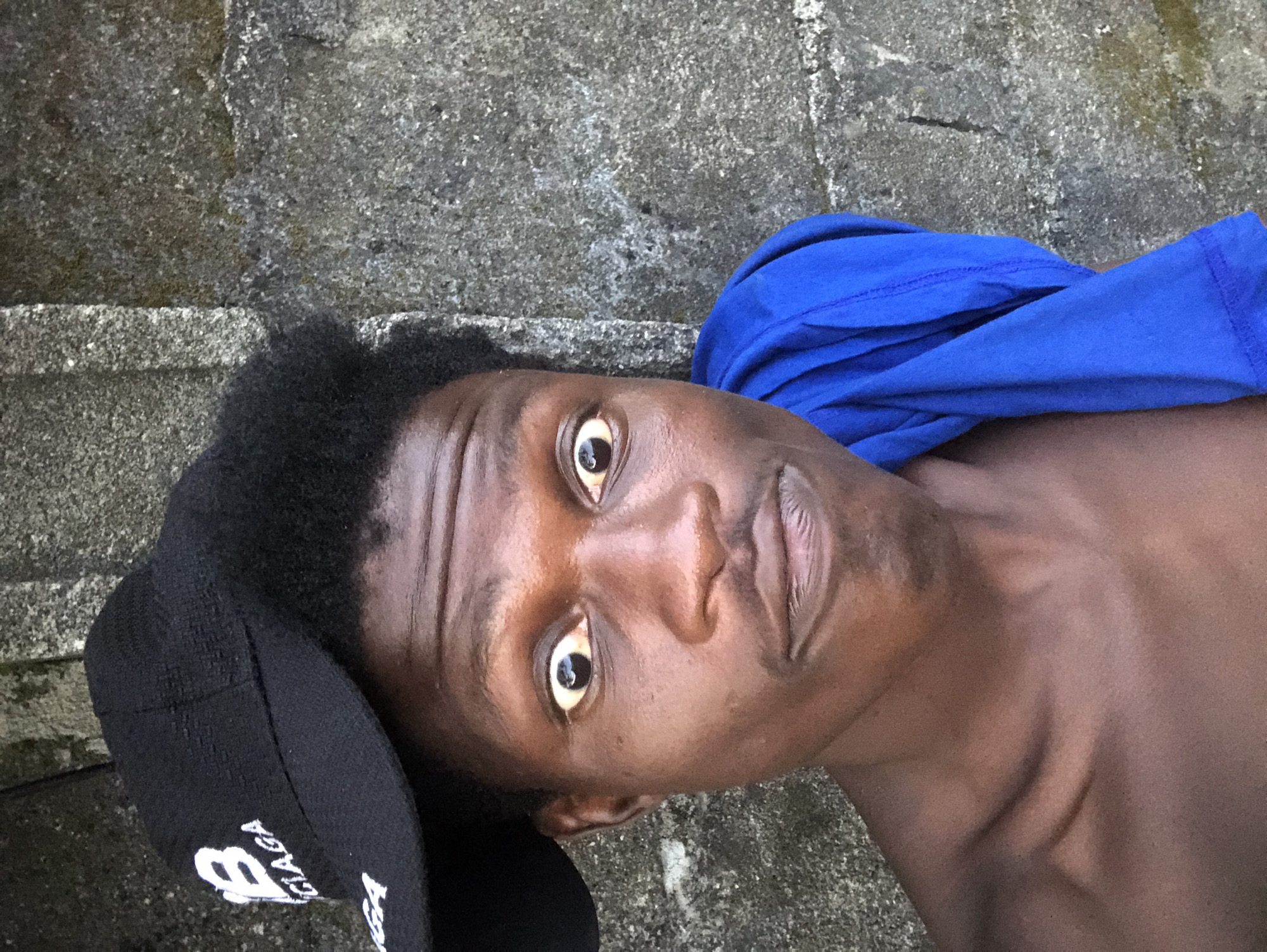 A dark-skinned male with an afro hairstyle is posing for a selfie in front of a concrete wall in Ado, Nigeria. He is wearing a black baseball cap with a white "B" logo and the word "CIAGA" on it, and a bright blue garment draped over his left shoulder. His eyes are wide open, and his mouth is slightly pursed, giving him an exaggerated expression. The concrete wall behind him appears weathered and textured, with some areas showing signs of moss or lichen growth. The lighting suggests it is daytime, with natural light illuminating the scene.