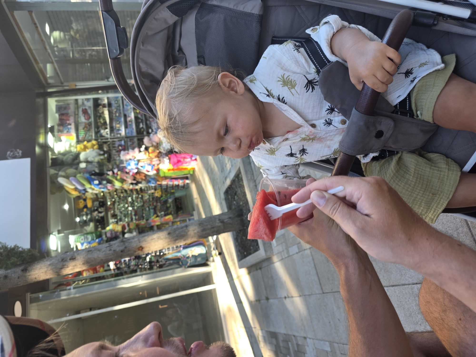 A man is feeding a young child watermelon in a stroller. The child is wearing a white shirt with a palm tree print and green shorts. The stroller is brown and gray. The scene is outdoors, with a shop in the background selling souvenirs. The ground is paved with stone. The image appears to be taken in the city of Sirius, Russia.