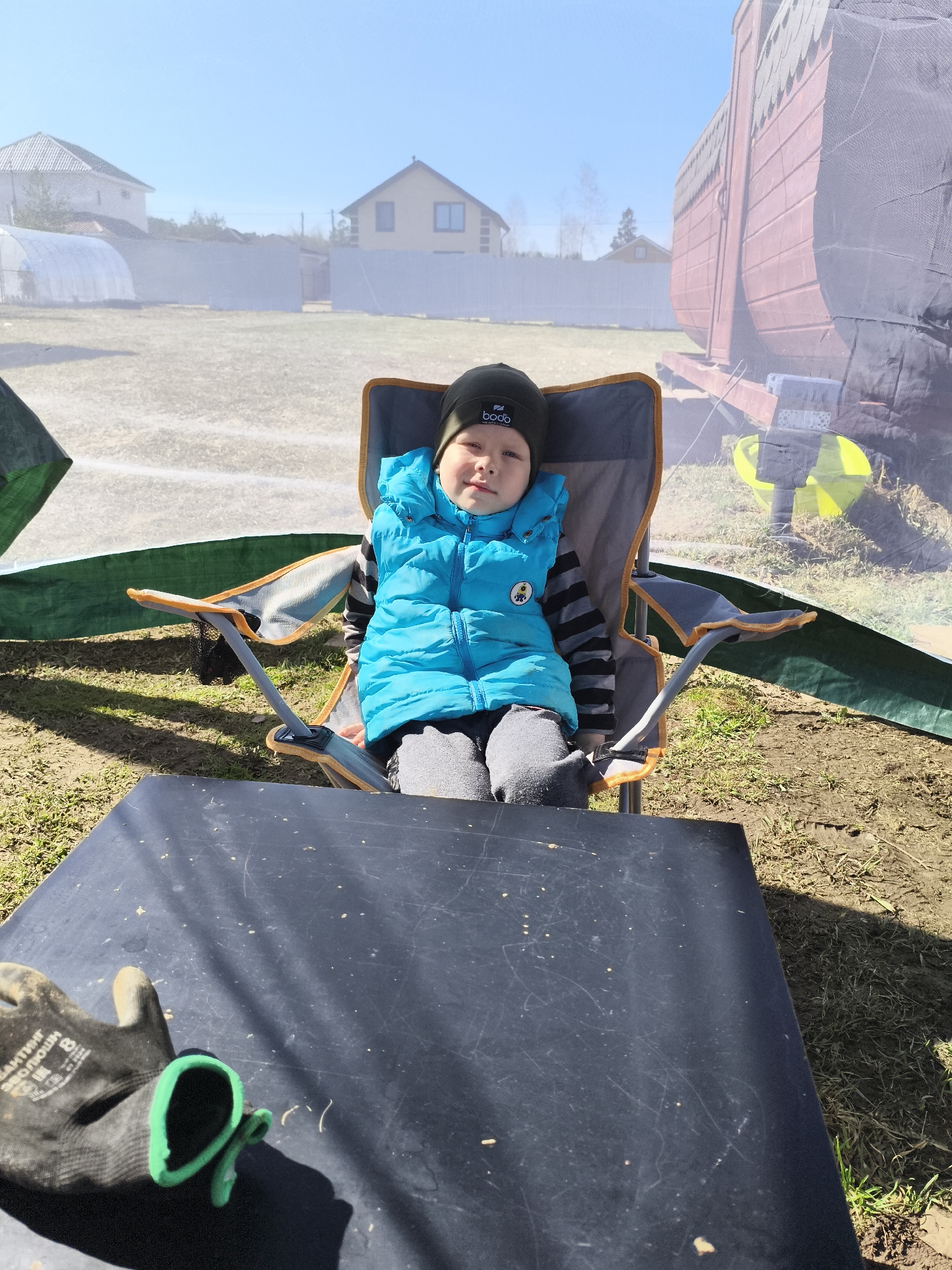 A young child wearing a black beanie, a bright blue vest, and a striped shirt sits in a camping chair outside. The chair is gray with orange trim. A black table is in the foreground with a work glove placed on it. The background features a mesh-like tent structure, and beyond the tent, there are houses and a greenhouse. The scene appears to be in a yard or outdoor area with grass and dirt. The location is Русский Вожой, Russia.