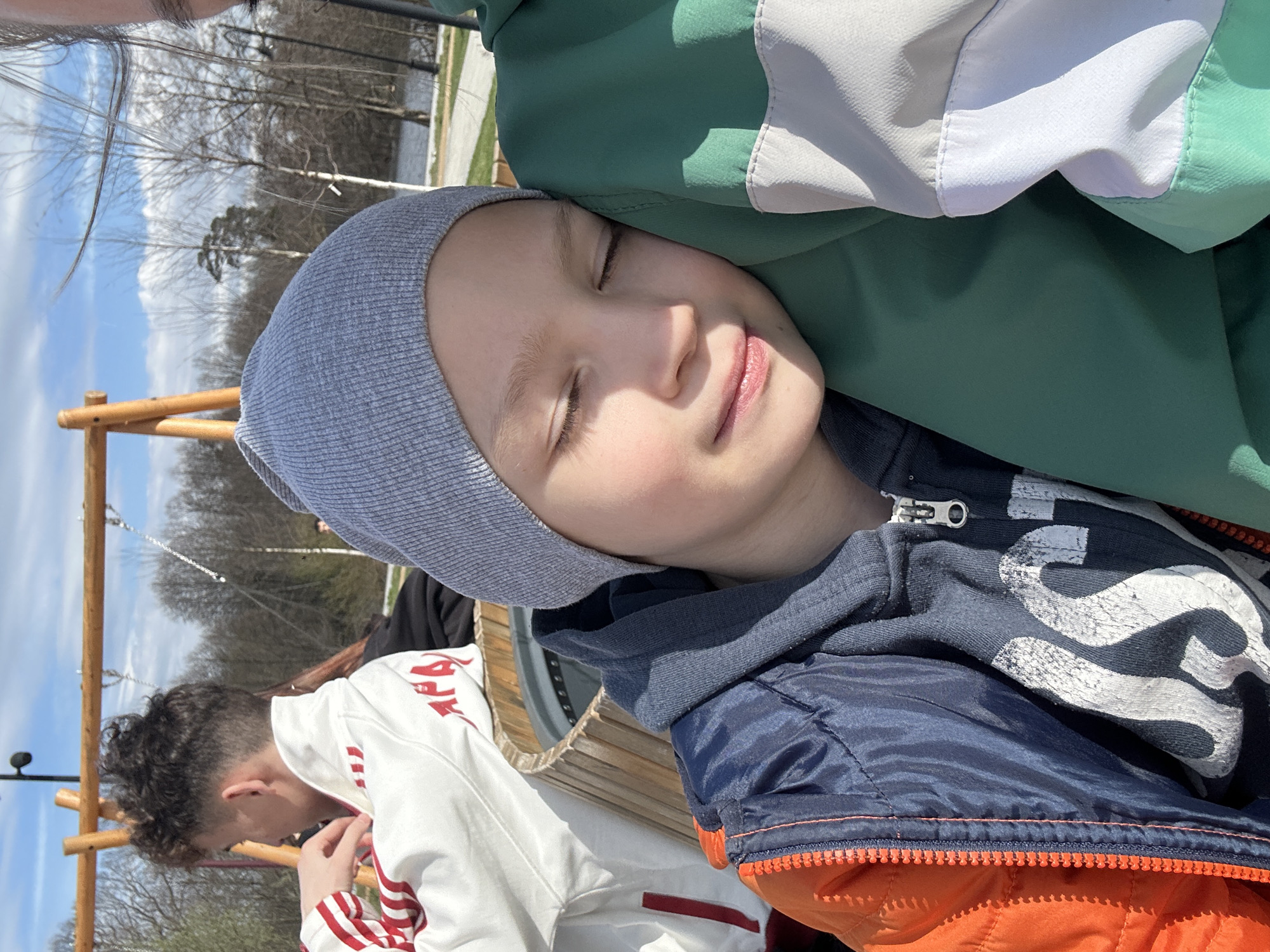 A young person with a gray knit cap and eyes closed rests their head against a person wearing a green and white jacket. The individual is wearing a zip-up hoodie with a navy blue and orange jacket on top. In the background, a person in a white and red jacket sits at a wooden structure with trees and a body of water visible. The location may be outdoors, as suggested by the sky and trees. The image was taken in Fryazino, Russia.