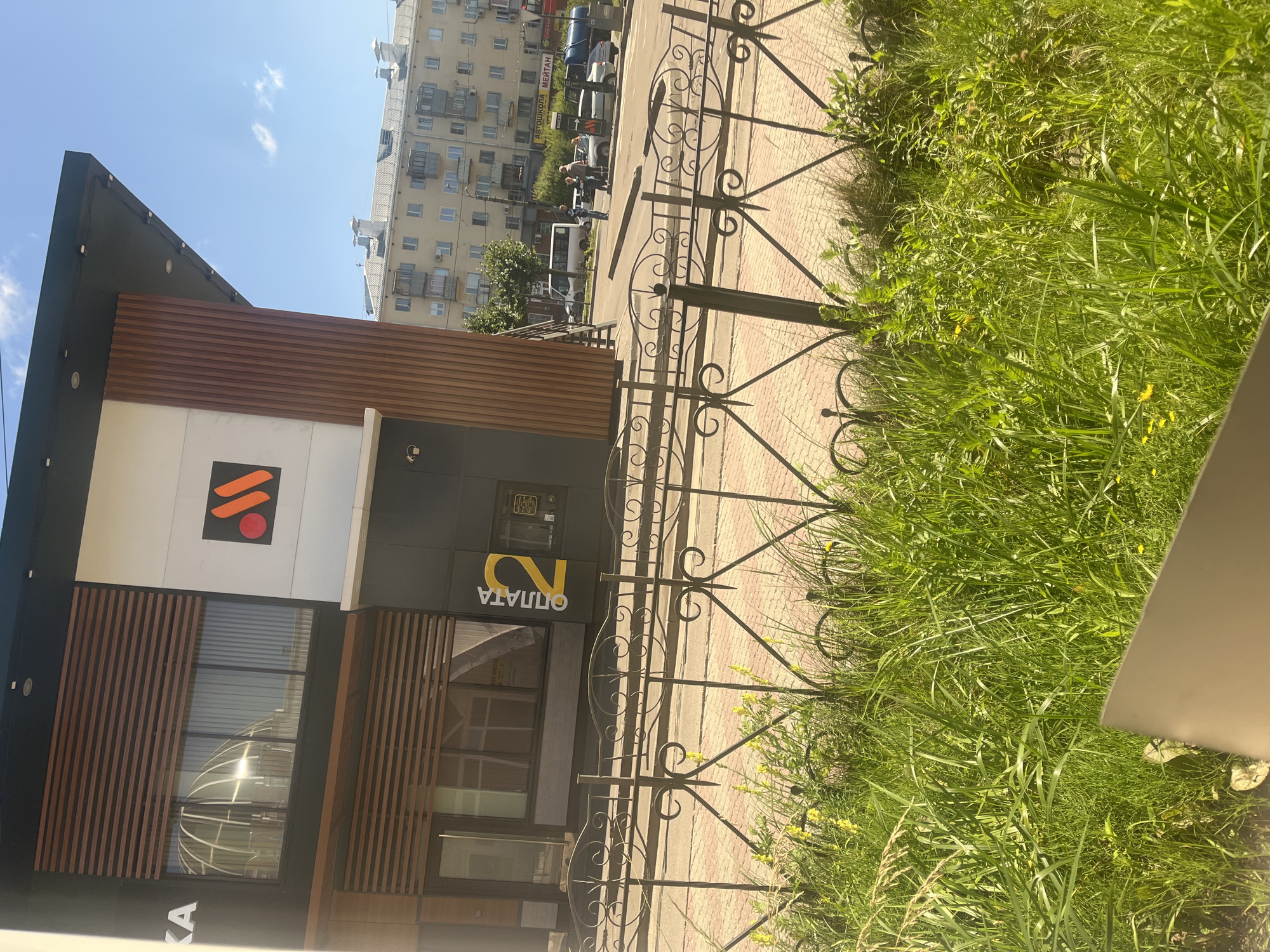 A modern, multi-story building with a dark facade and wooden paneling stands on a sunny day. The building has large windows with wooden blinds and features a prominent logo with an orange "M" on a white background. To the right of the logo, the word "ОПЛАТА" (payment) and the number "2" are displayed in yellow on a dark gray section of the building. In front of the building, a decorative metal fence borders a sidewalk paved with red bricks. Beyond the sidewalk, a street with cars and pedestrians is visible, leading to apartment buildings in the background. Lush green grass and yellow wildflowers grow in front of the fence, suggesting a park-like or roadside setting. The scene appears to be in Ivanovo, Russia, as evidenced by the architectural style and Cyrillic signage.