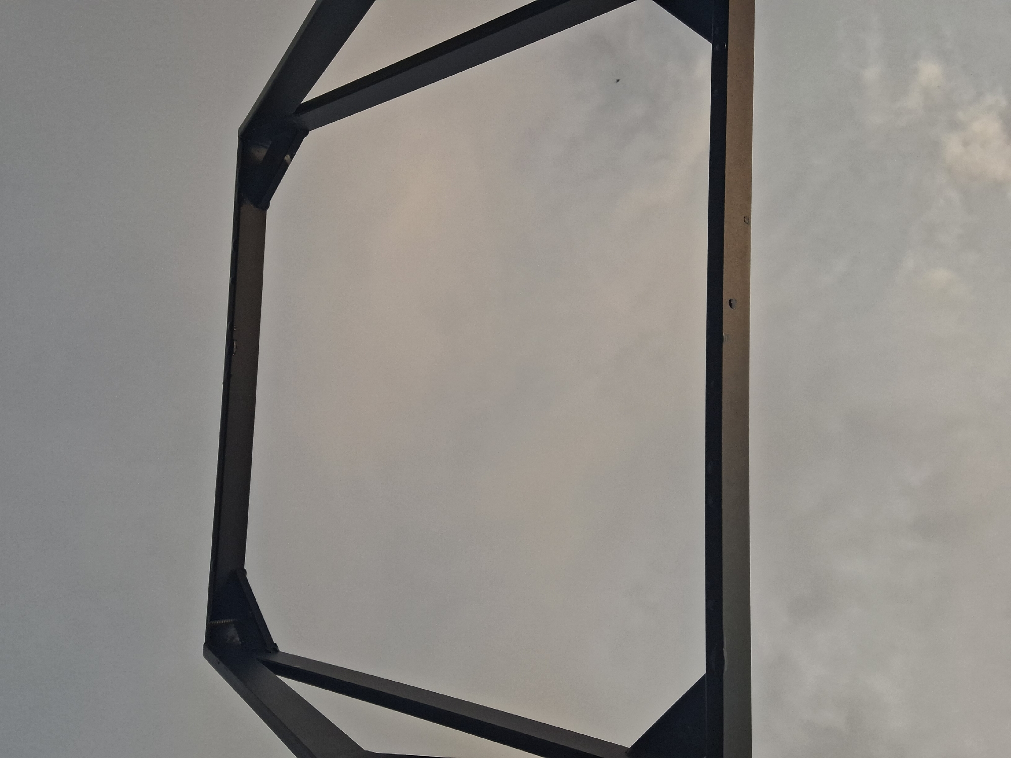 A low-angle shot depicts a rectangular metal frame structure against a cloudy sky. The frame consists of dark, possibly black, metal beams. The frame's construction includes diagonal support beams at the top and bottom corners. The sky visible through the frame is overcast with varying shades of gray. No people, specific objects, or actions are immediately discernible. The location and environment are indeterminate based on this visual information alone; the reference to Anipemza, Armenia is contextually unverified within the image itself.