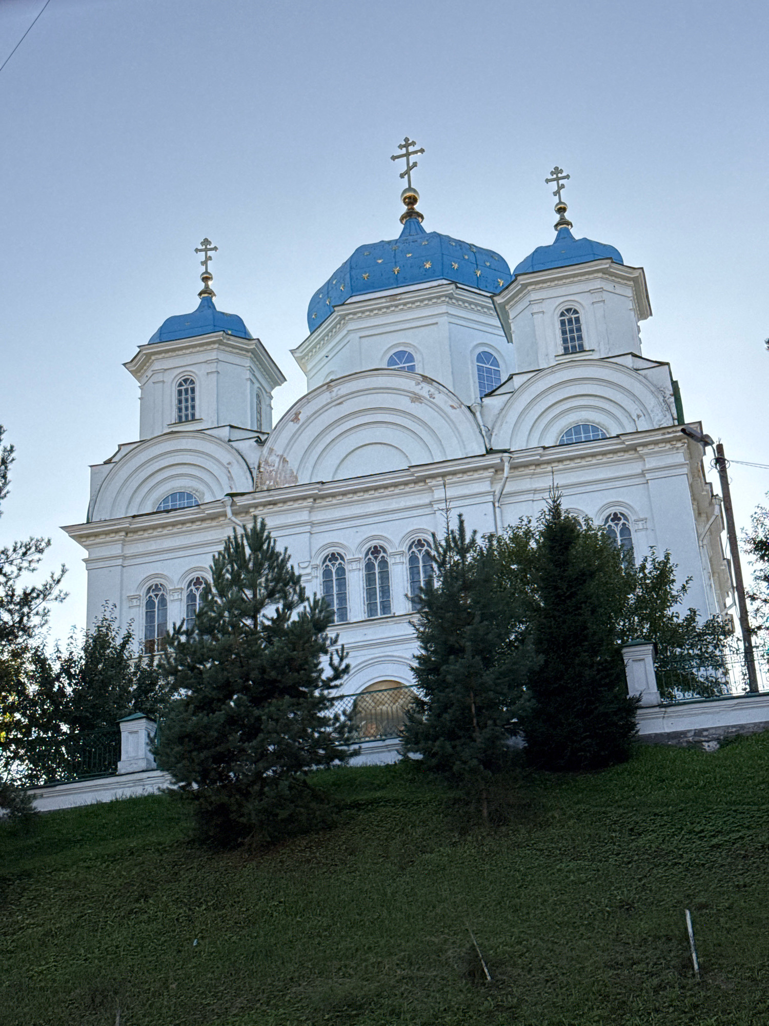 The image shows the Borisoglebsky Monastery in Torzhok, Russia. The monastery is a large, white building with blue domes topped with crosses. It is situated on a grassy hill, with trees in the foreground. The sky is clear and blue. The building exhibits a classical architectural style with multiple levels, arched windows, and decorative elements.