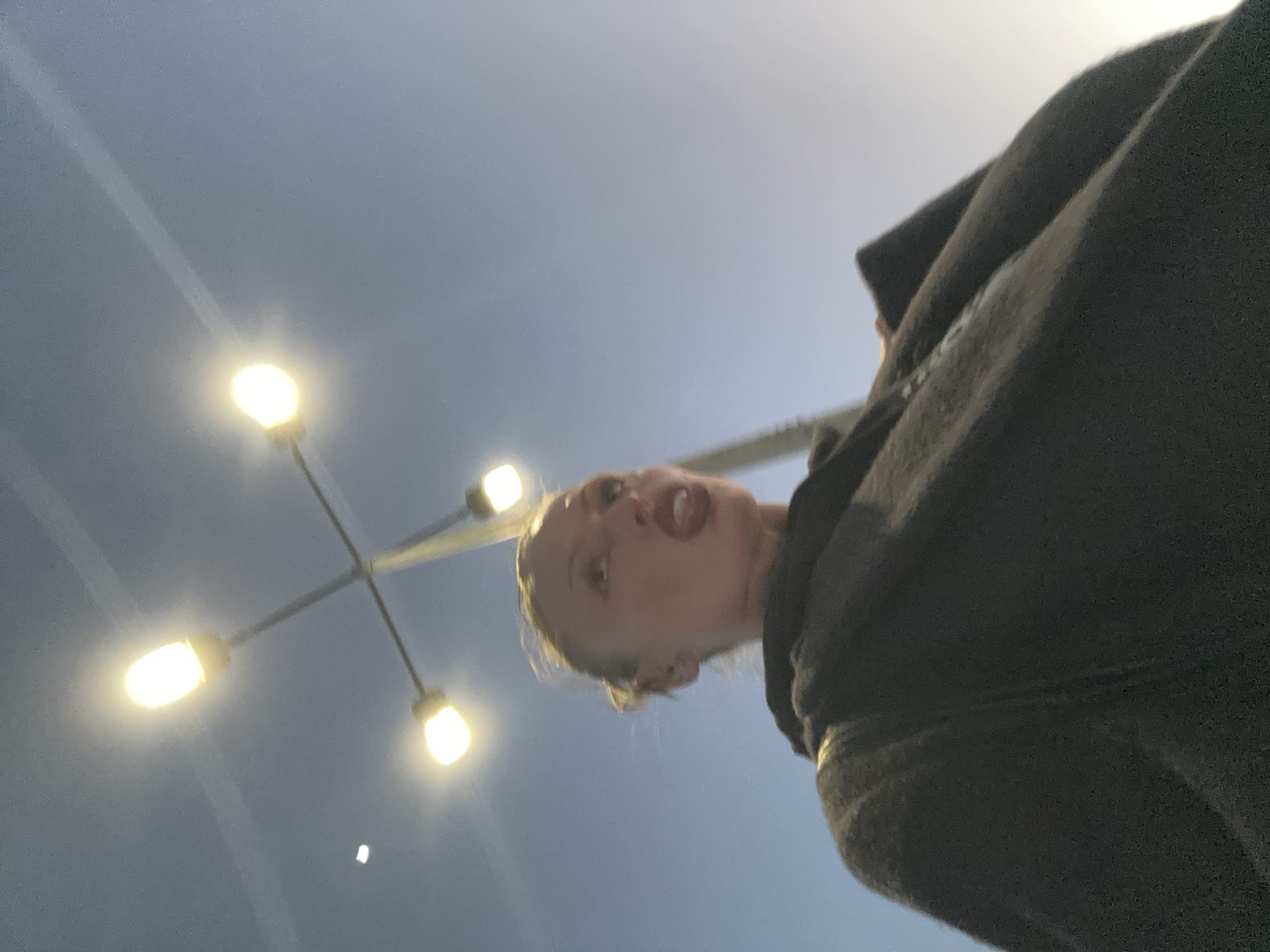 A low-angle photograph, captured in Sevenoaks, United Kingdom, depicts a female subject looking upwards towards four illuminated streetlights against a twilight sky. The subject wears a dark-colored overcoat, has light hair pulled back, and her mouth is open with her tongue slightly visible. Her gaze is directed towards the bright, oval-shaped streetlights, which are mounted in a cross formation on a single pole. The sky is clear, displaying a light blue-grey hue, with a faint white point of light in the upper left. The scene's illumination is primarily from the artificial streetlights, indicating dusk or nighttime.