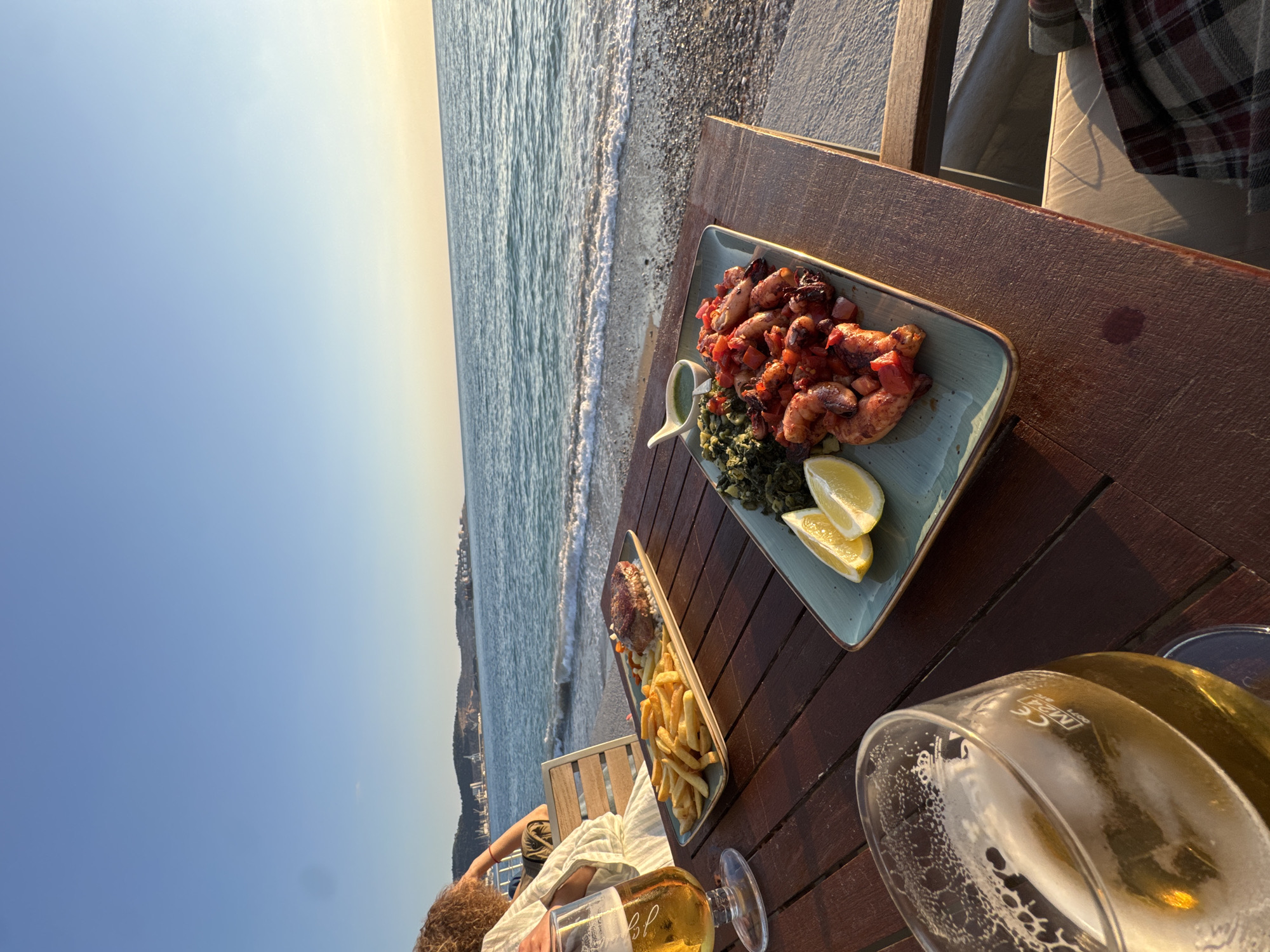The scene is set at a restaurant on the beach in Bar, Montenegro. It's likely either daytime or dusk, judging by the sky and light. A wooden table is in the foreground, and on it are two plates of food and two glasses of beer. One plate appears to have grilled octopus with vegetables and lemon slices. The other plate contains a steak and french fries. In the lower-left corner of the frame, a portion of a person can be seen sitting at the table. The beach and the sea are visible in the background, with buildings and hills on the horizon. The image is taken at an angle, giving a broader view of the seaside environment.