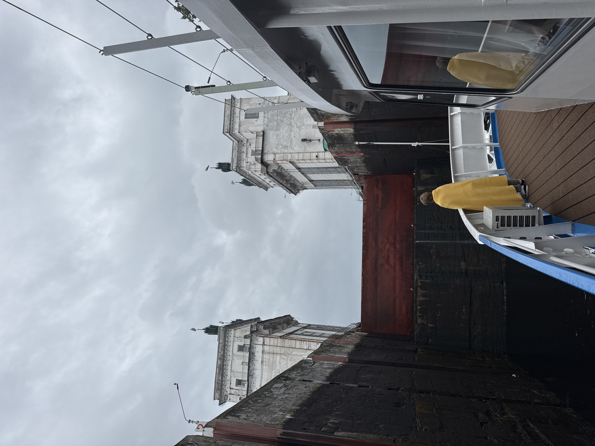 The image captures a view from a boat as it enters a lock in Podporozhye, Russia. The boat, partially visible on the right, has a white exterior and a brown wooden deck. A person is standing on the deck, wrapped in a bright yellow blanket, looking towards the lock gates. The lock itself is a formidable structure with high, dark walls, and a large red gate in the distance. Two imposing white buildings with ornate architecture and statues on top flank the lock entrance. The sky is overcast, contributing to a somewhat somber atmosphere.