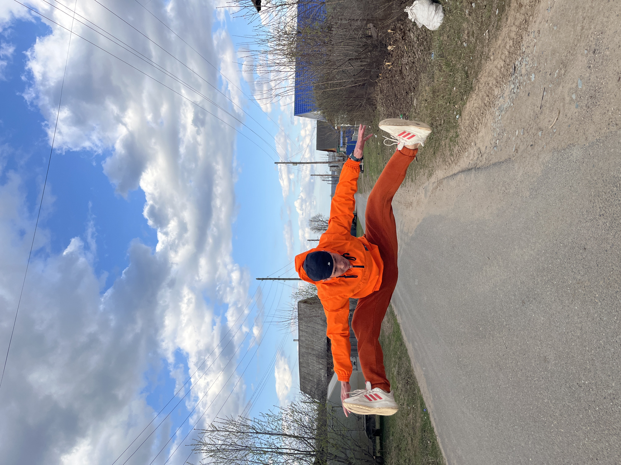 An outdoor, rotated image taken in Chaikovsky, Russia, features a person performing a dynamic high kick or split. The individual, likely male, is dressed in a vibrant orange hooded jacket, matching orange pants, and white sneakers adorned with red Adidas stripes. They are captured in mid-action with one leg extended high into the air, while their upper body leans forward.
The setting is a rural or semi-rural area under a bright, partly cloudy daytime sky, showcasing large white and gray clouds interspersed with patches of blue. In the background, simple houses or structures are visible, some featuring distinctive blue roofs. Bare trees and sparse bushes line the edges of the scene, suggesting either early spring or late autumn. Power lines stretch horizontally across the sky. The ground beneath the person consists of an asphalt road that transitions into a dirt and gravel shoulder, with patches of grass and a white bag on the embankment. No specific emotions are discernible, and the notable text is the "Adidas" logo on the sneakers.