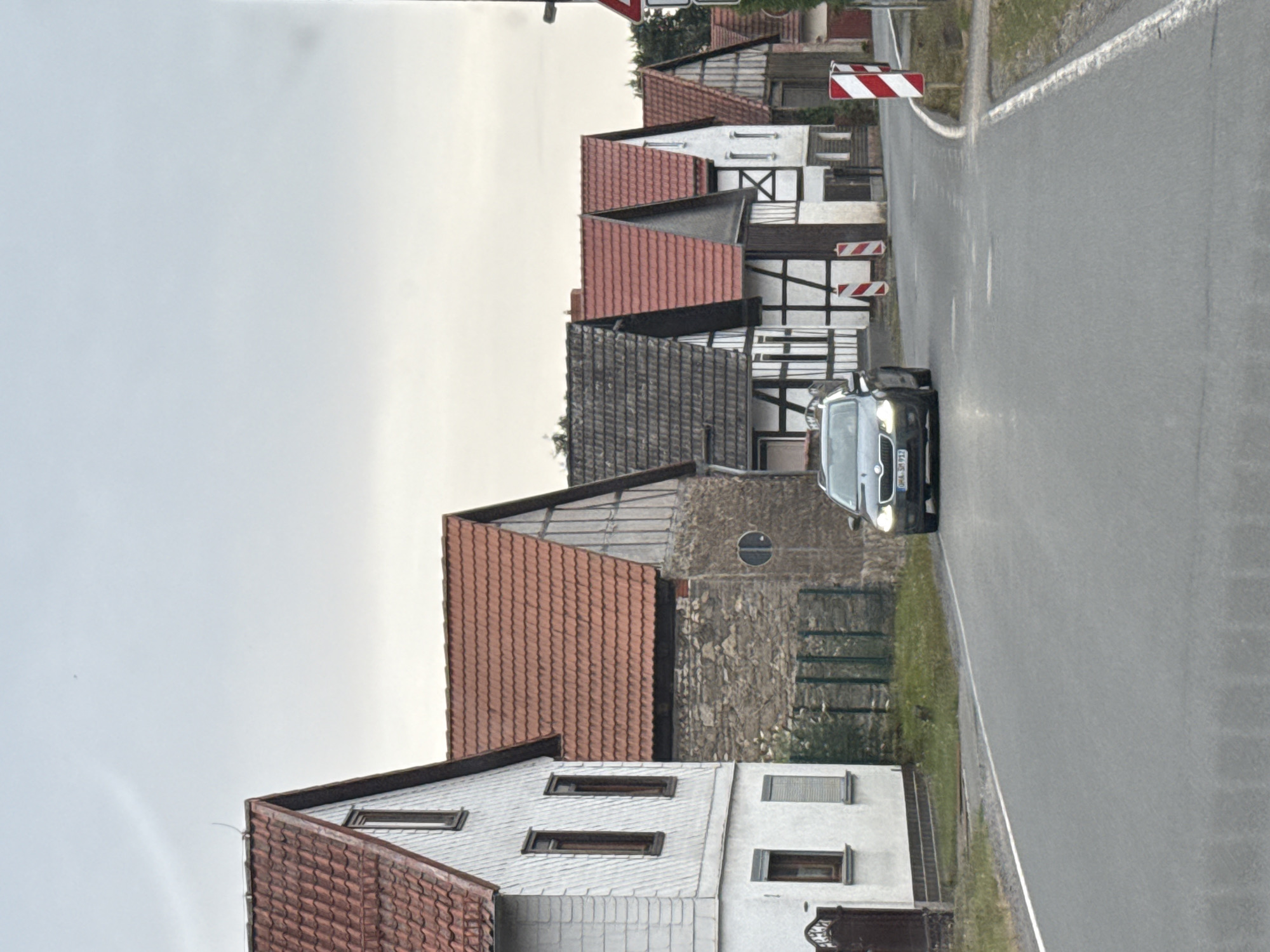 The image depicts a street scene in the city of Hohenstein, Germany. A dark-colored Skoda car is driving on a paved road in the foreground, with its headlights illuminated. The road has a white line marking the edge on the right side. To the left of the road are several buildings with traditional German architecture, featuring red-tiled roofs and white-washed facades. Some of these buildings have exposed timber framing. In front of one of the buildings, a dark metal gate is visible. A green metal fence runs along the property line. Beyond the buildings, trees are present. On the right side of the road, there are two red and white striped traffic posts and a red and white triangular sign with a black symbol, indicating a hazard or warning. The sky is overcast and light grey. The overall scene suggests a village or rural setting.