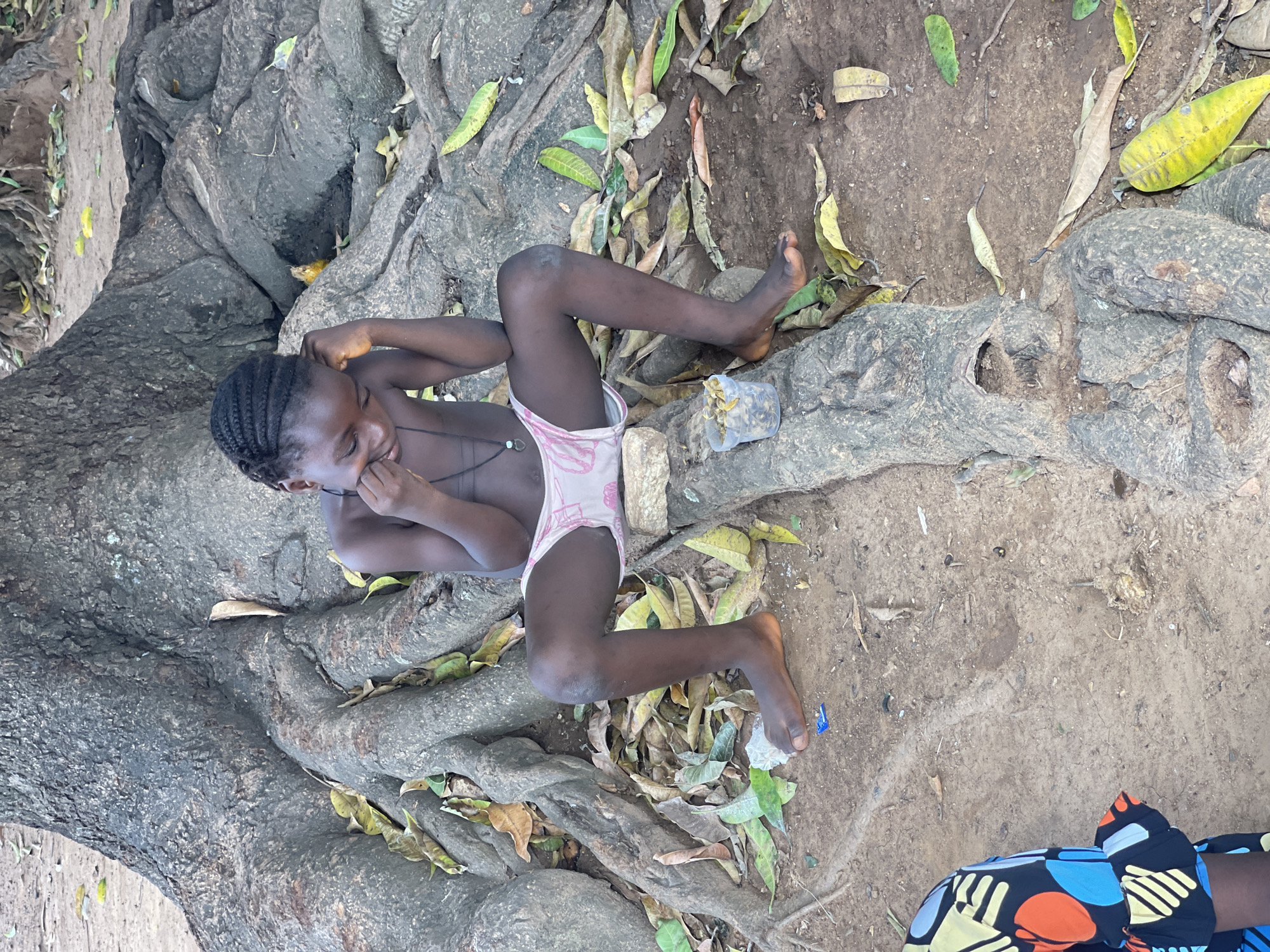 A young, dark-skinned child lies on the exposed roots of a large tree. The child is facing left with their head resting on the roots and legs splayed outward. The child is wearing pink patterned underwear and a necklace with a pendant. One hand is near their face, and the other is bent at the elbow. A clear plastic cup sits near the child’s leg. Yellow and brown leaves are scattered around the tree roots and on the ground. The ground appears to be dirt. A person wearing a brightly colored shirt with blue, orange, and yellow patterns is partially visible in the lower right corner of the frame. The location is in Akwanga, Nigeria.