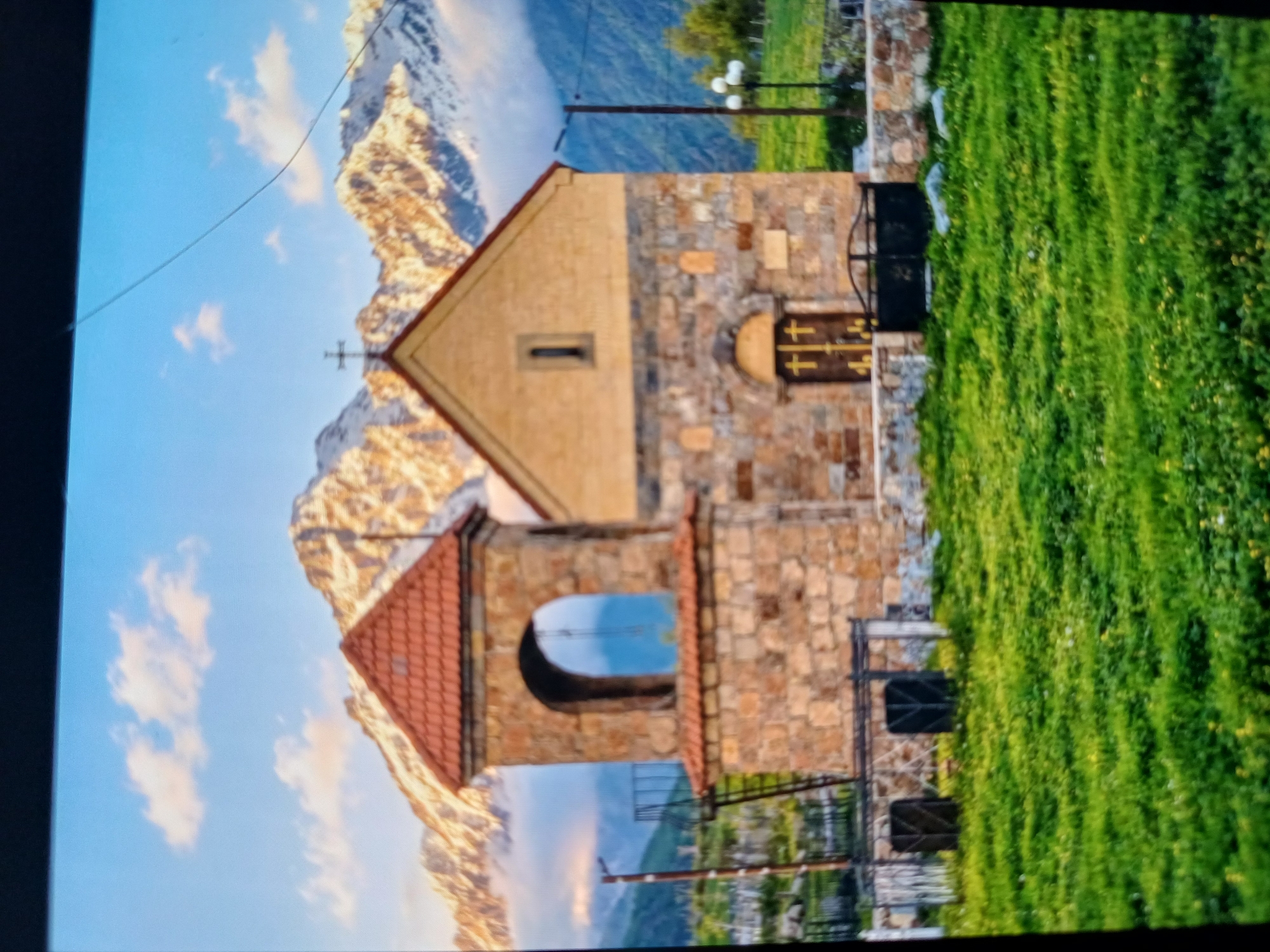 The image shows a small stone church with a bell tower, situated in a grassy field. Snow-capped mountains form the backdrop under a blue sky with scattered clouds. The church features a red-tiled roof, stone walls, and a wooden door with cross motifs. A metal gate is in front of the church, and a lantern is visible on a pole to the right. The bell tower has an arched opening. The scene appears to be in the city of Stepantsminda, Georgia, given the mountainous terrain.