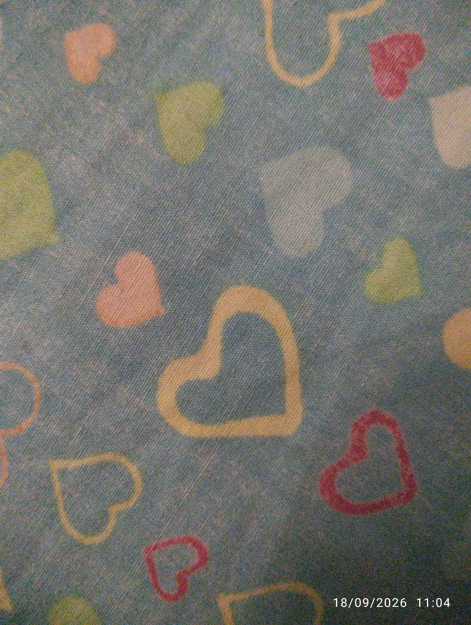 The image is a close-up, overhead shot of a light blue fabric with a pattern of colorful hearts. The hearts are in various shades of pink, yellow, and green, and some are outlined in a thicker yellow line. The fabric appears to have a textured, woven surface. There is a timestamp in the bottom right corner that reads "18/09/2026 11:04". The image does not provide any information about the city of Chattogram, Bangladesh, or any specific location within it.