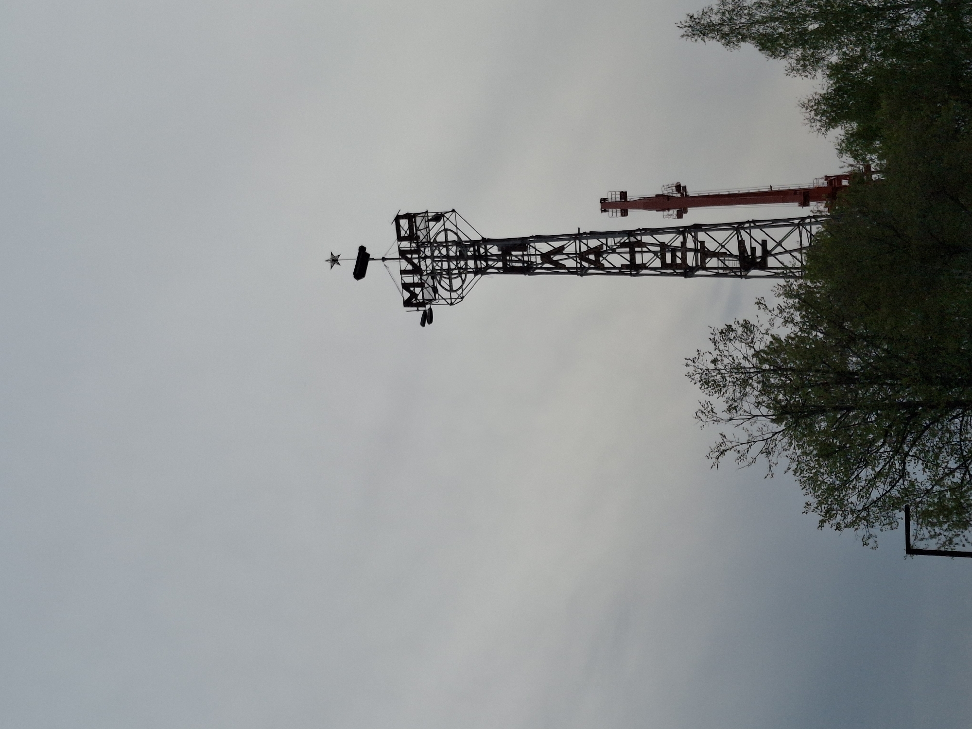 The image shows a telecommunications tower in Chaikovsky, Russia. The tower is constructed of lattice steel, painted black, and has a star-shaped decoration on top. Another red crane is visible in the background. Trees are to the right. The sky is overcast and gray.