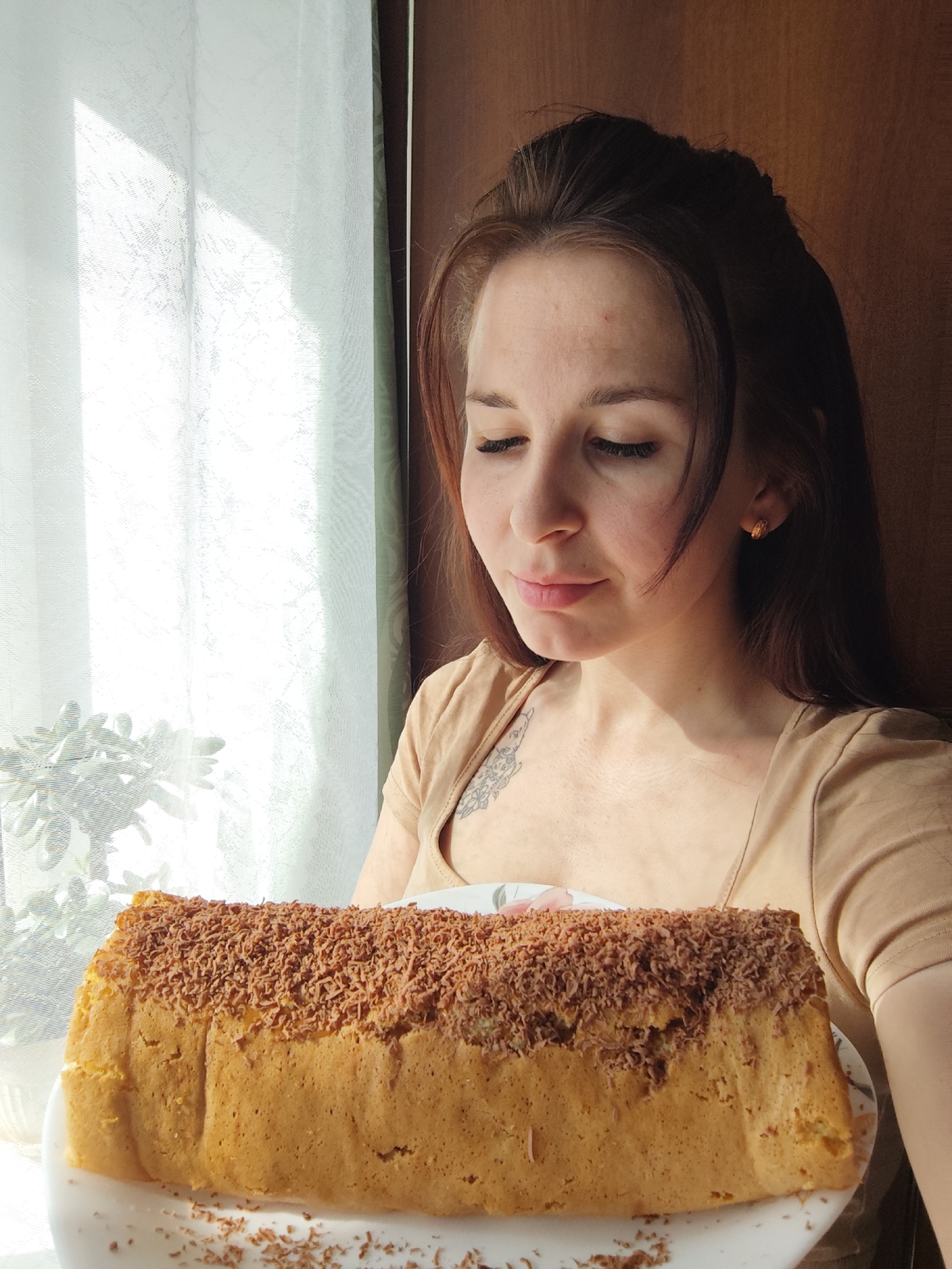 A woman with long brown hair and fair skin holds a rectangular cake on a white plate. The cake is golden brown and covered in chocolate shavings. The woman is wearing a tan t-shirt and earrings. She has a tattoo visible on her upper chest. A window with sheer curtains and a potted plant is behind the woman to the left. A dark wooden wall is behind her to the right. The location appears to be indoors. The city of Фоки, Russia, is not discernible from the image.