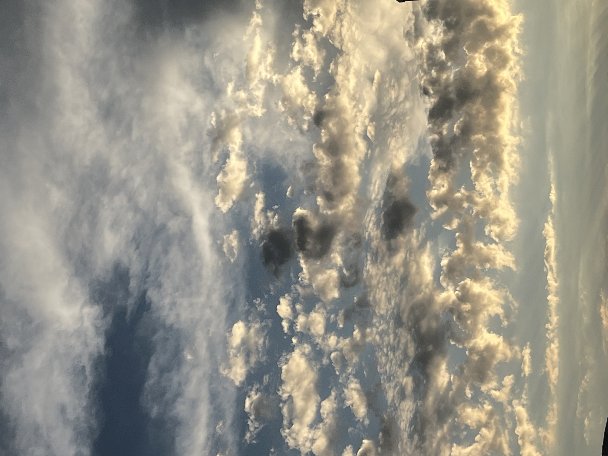 The image displays a dramatic sky filled with a mixture of clouds, suggesting a dynamic weather system. The clouds vary in texture and color, with some appearing bright white and illuminated by sunlight, while others are darker and more shadowed, indicating depth and possibly approaching precipitation. Wispy, thin clouds stretch across the upper portion of the frame, contrasting with the more voluminous, cumulus-like formations below. The lighting suggests it could be either sunrise or sunset, given the warm, golden hues on some of the clouds, though the overall color palette also includes cooler blues and grays of the sky. There are no people or man-made objects visible, focusing solely on the natural phenomenon of the sky. The setting appears to be outdoors, under an open sky. The image is oriented vertically, making it a portrait shot of the sky. The weather appears to be mixed, with clear sky visible in some areas and dense cloud cover in others. No text is visible.