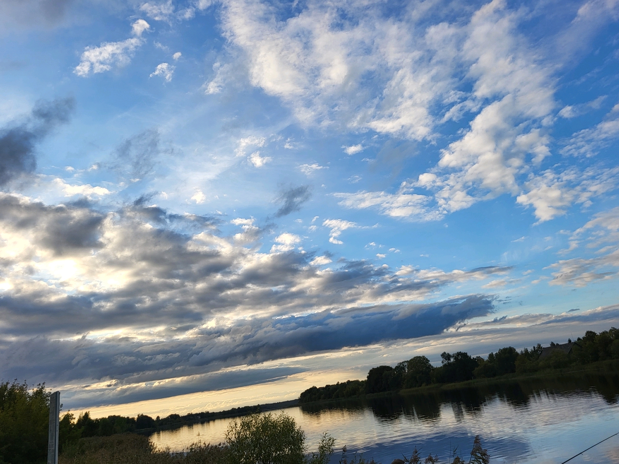 The image captures a wide-angle view of a riverbank and the sky, likely taken in Kalnciems, Latvia, during the evening. The sky is dominated by a dramatic mix of clouds, with patches of bright blue interspersed with darker, more ominous formations. The sun appears to be setting or has recently set, casting a warm, golden light across the scene, particularly on the water and the underside of some clouds.
The foreground on the left side features dense vegetation, including trees and reeds, with a single tall, weathered wooden post visible. The river occupies the mid-ground, reflecting the sky and the tree line that stretches along the opposite bank. The opposite bank is lined with a dense forest of trees, with some hints of structures or buildings nestled within the foliage. The water is calm, with gentle ripples disturbing the reflection. The overall atmosphere is serene and natural.