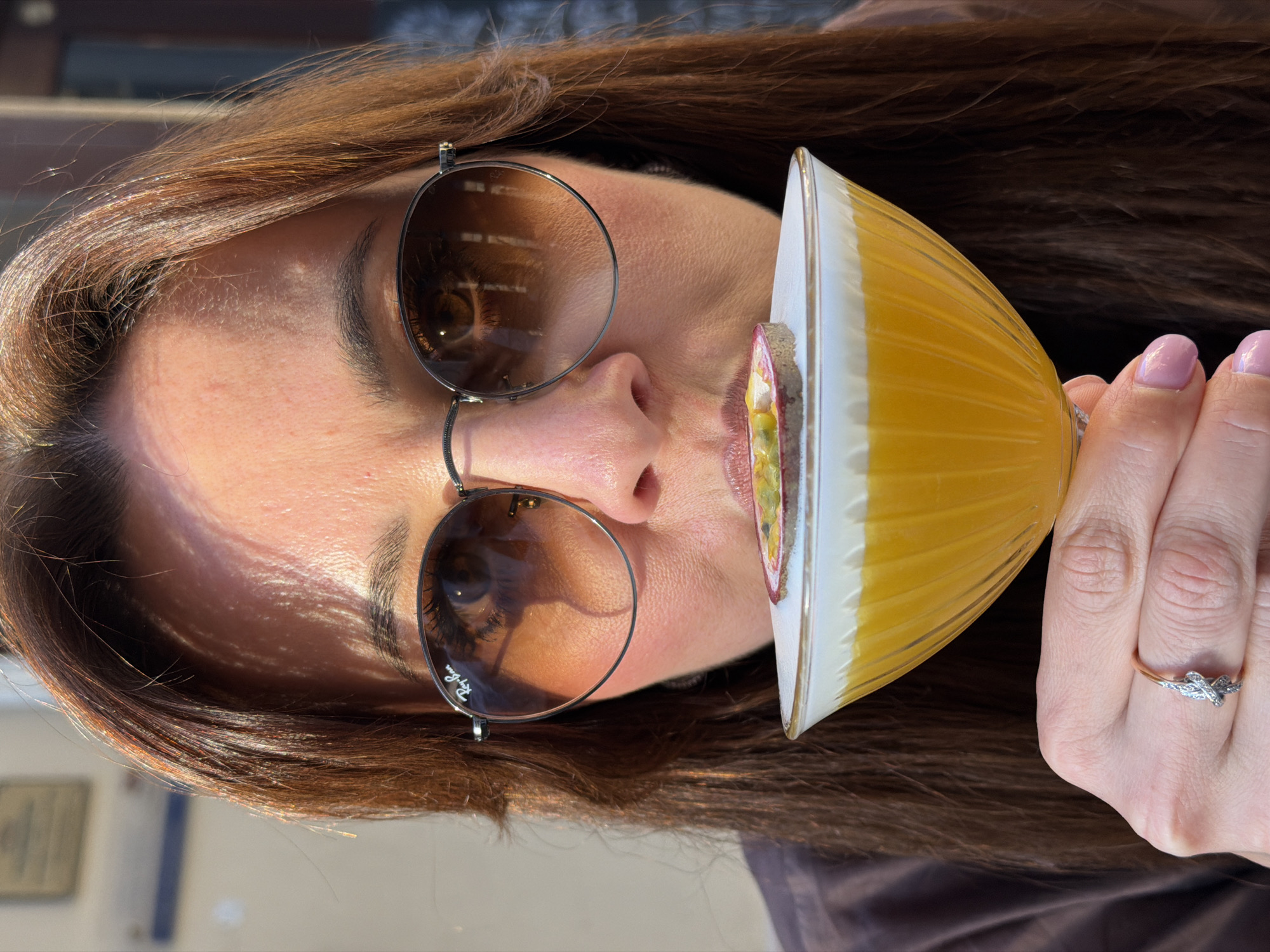 A close-up, horizontally oriented photograph captures a woman enjoying a cocktail outdoors in Riga, Latvia. The bright, sunny daytime lighting emphasizes the auburn highlights in her hair and skin. She wears round, dark-tinted sunglasses with metal frames, and the "Ray-Ban" logo is clearly visible on the left lens, partially obscuring her eye.
Her left hand, adorned with a silver-colored ring featuring a knot or bow design on her ring finger and light pink polished nails, holds a distinct cocktail glass. The glass is a ribbed or fluted coupe style, yellow with a white rim, containing a yellowish beverage. A slice of passion fruit, showcasing its seeds and pulp, garnishes the rim of the glass, positioned near her slightly parted lips as if she is about to drink. The background is softly blurred, suggesting an outdoor setting like a cafe or patio, contributing to a relaxed ambiance.