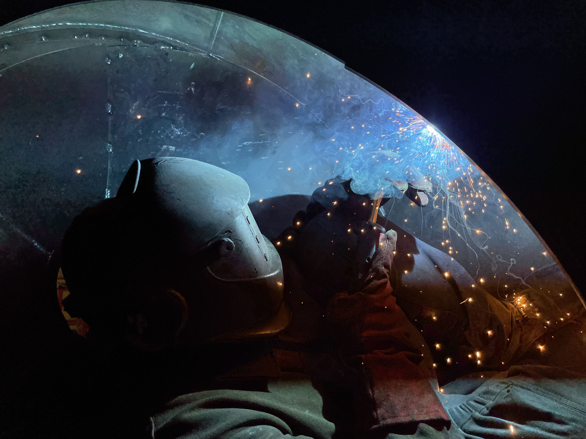A welder is captured mid-operation, enveloped in protective gear consisting of a helmet and gloves, amidst a shower of sparks and smoke emanating from the welding activity. The environment appears to be a dark, enclosed workspace. There is no information to suggest that the image was taken in Čeluga, Montenegro.