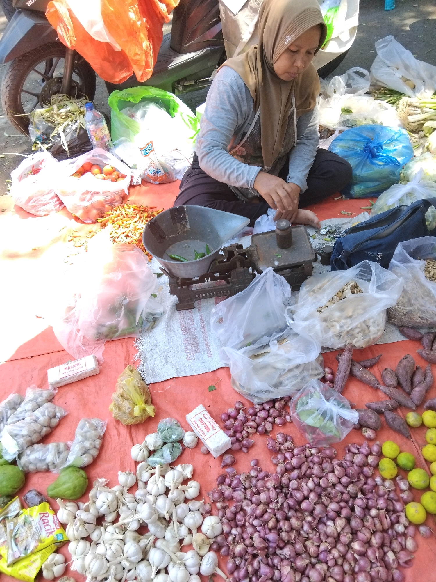 A woman wearing a light brown hijab and grey long-sleeved shirt is sitting on a reddish-orange tarp at a street market in Kepuh Kiriman, Indonesia. She is focused on weighing produce using a vintage scale. Various food items are displayed around her on the tarp, including piles of red onions, garlic bulbs, sweet potatoes, green limes, and tomatoes. Several plastic bags containing produce are also visible, as are two white rectangular packages labeled "TEMPE MURNI MALANG". In the background, a motorcycle is parked, and more plastic bags of produce are stacked. The ground appears to be dirt or pavement. The lighting suggests it is daytime.