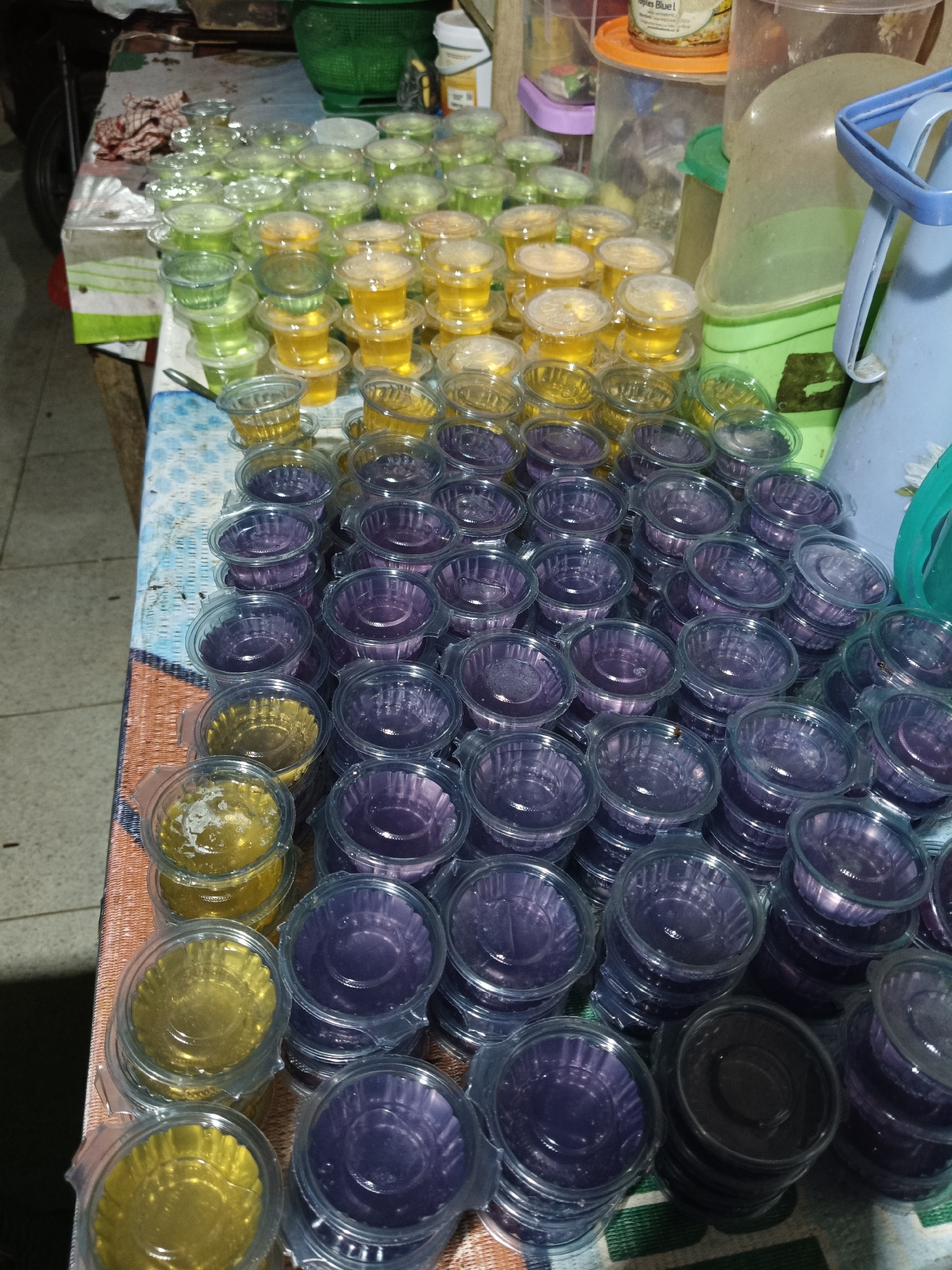 A street vendor in Rancaseneng, Indonesia, displays numerous small, clear plastic containers filled with jelly. The jellies are in two colors: yellow and purple. They are arranged in stacks on a table covered with a patterned cloth. In the background, larger containers and other items suggest a market or food stall setting.