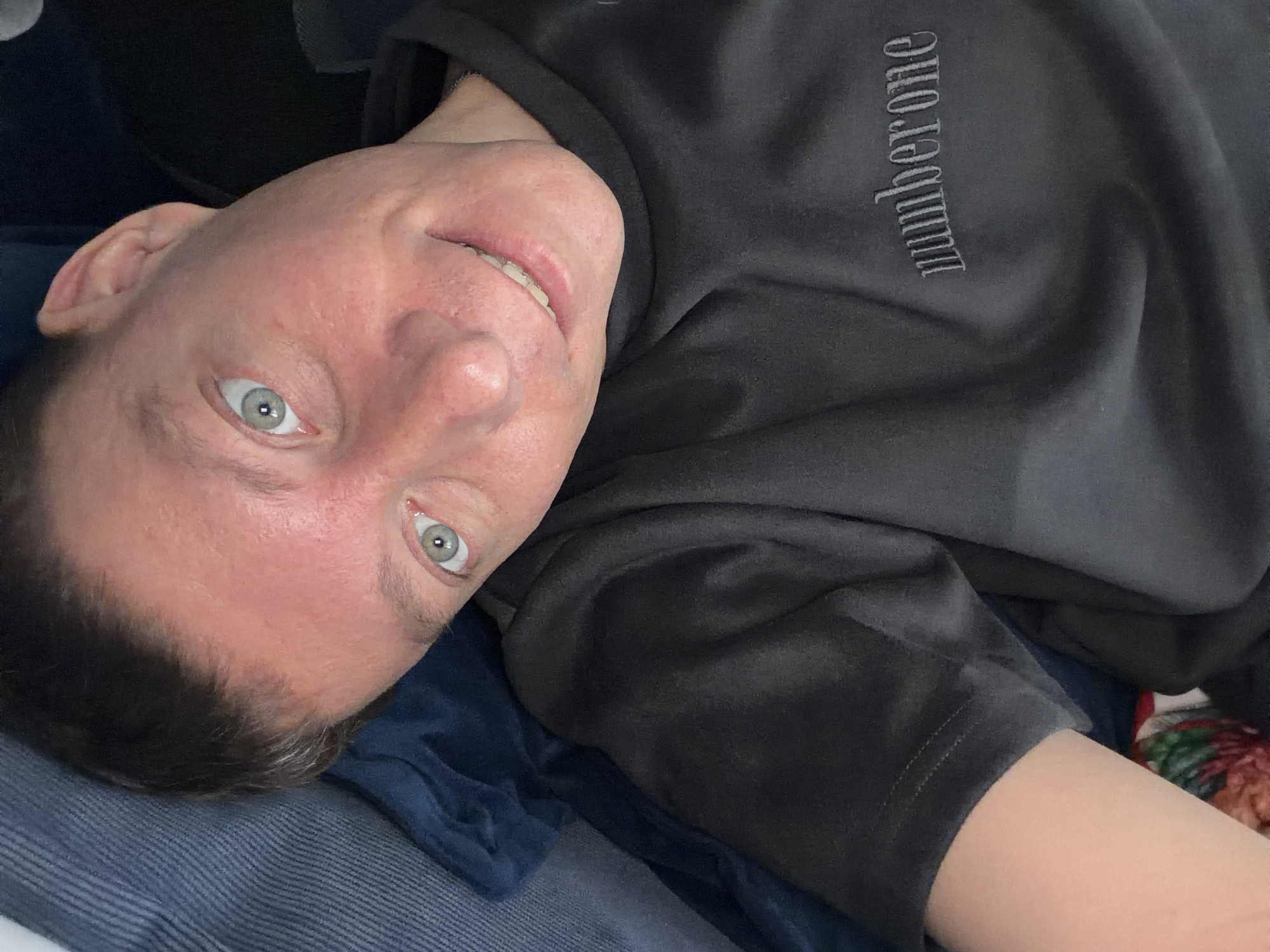 The image is a close-up, high-angle selfie of a person with light skin, light eyes, and short dark hair. The person is wearing a dark gray t-shirt with the word "numberone" embroidered in a darker gray thread across the chest. The person is lying down, with blue fabric visible behind their head and shoulder. The lighting suggests an indoor setting. There is no discernible location in Moscow, Russia.
