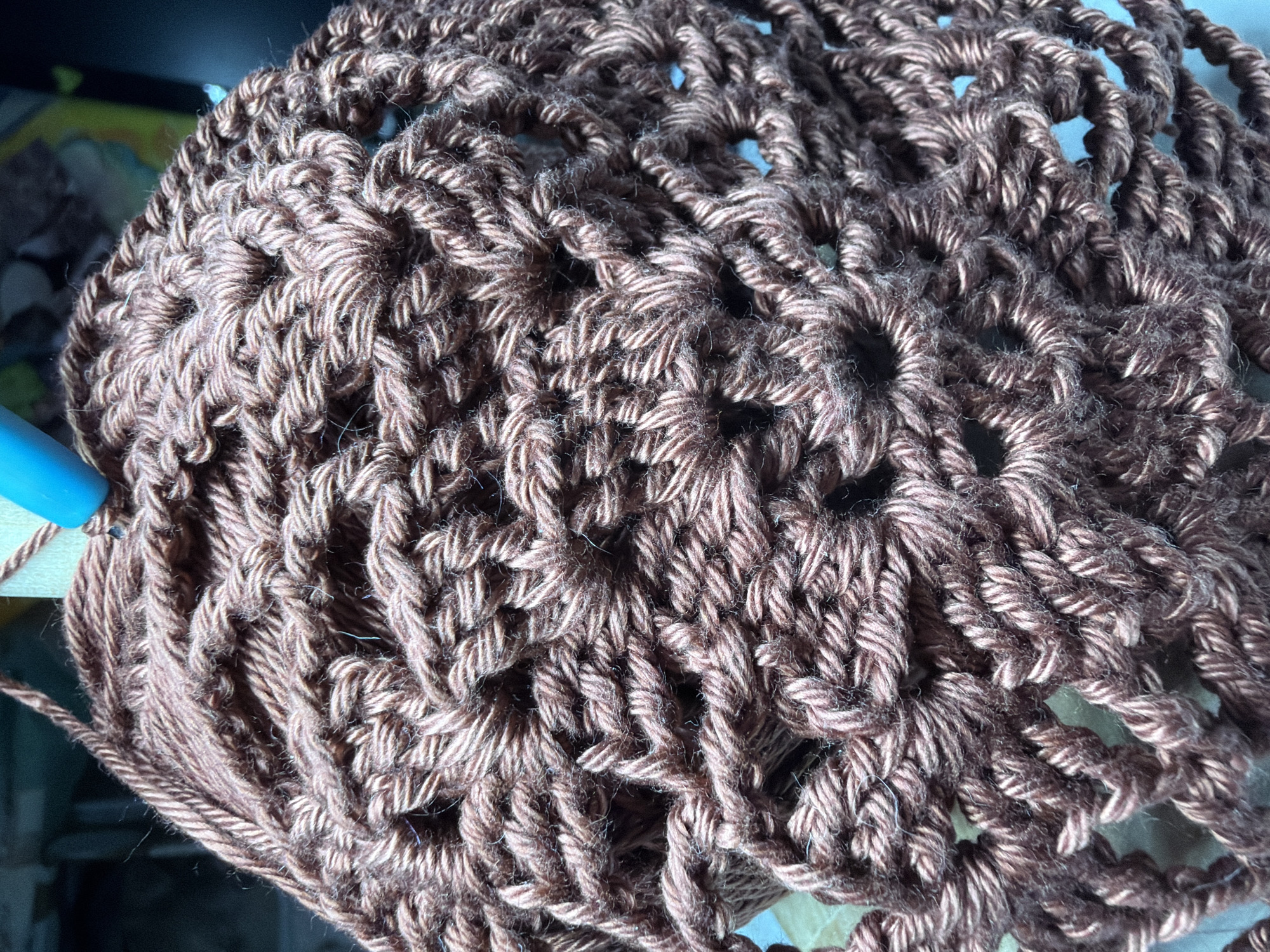 The image shows a close-up of a partially crocheted brown hat. A blue crochet hook is visible in the lower left corner, inserted into the yarn. The crochet pattern features dense stitches interspersed with circular open spaces. The yarn has a slightly fuzzy texture. The scene does not include any information that can be identified within the city of Chaikovsky, Russia.