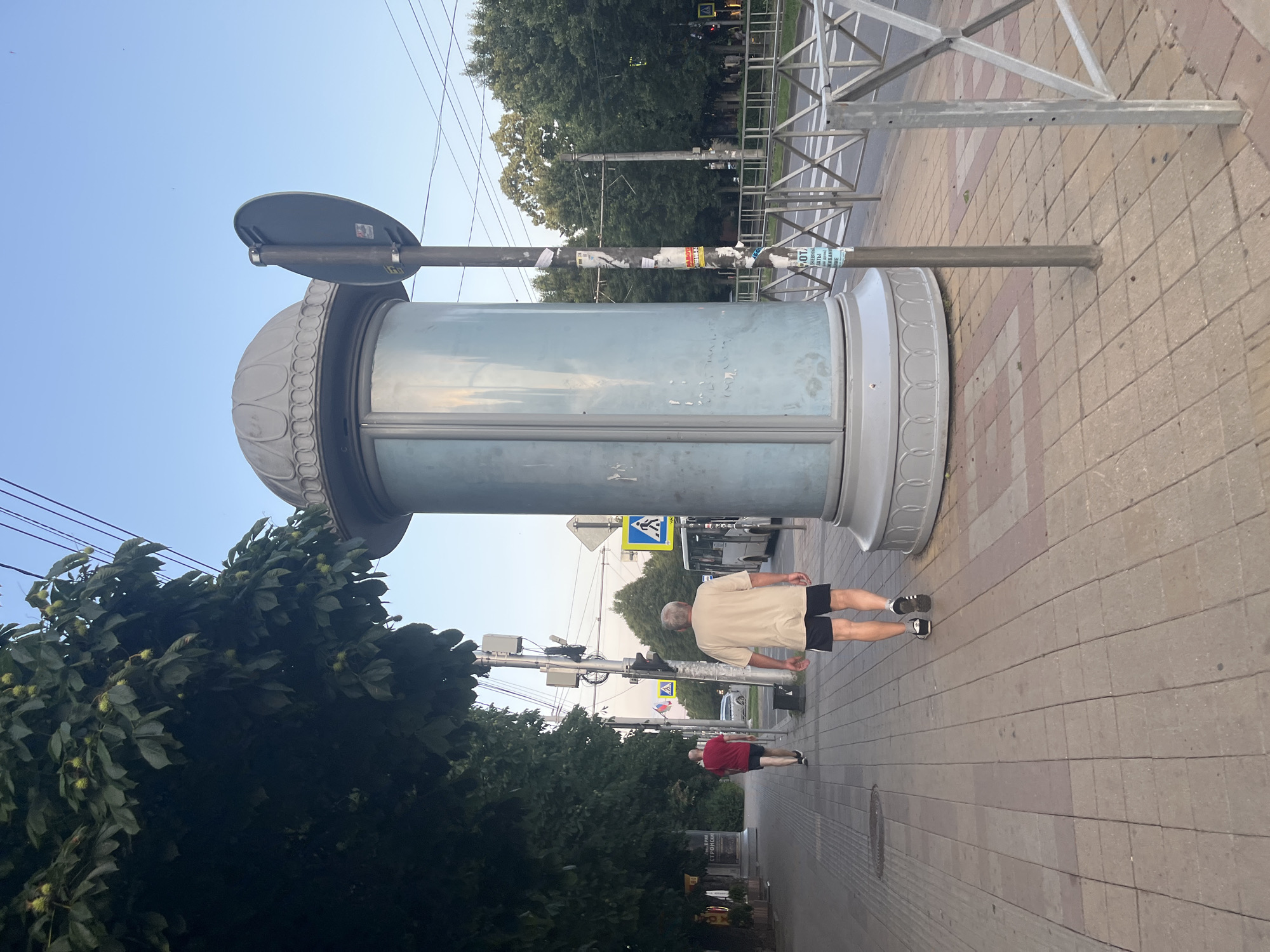 The image depicts a street scene in Krasnodar, Russia, on a clear day with a light blue sky. In the foreground, a large, cylindrical advertisement column with a decorative domed top stands on a sidewalk paved with red and beige rectangular tiles. A metal pole with various stickers is attached to the column, supporting a circular road sign. A metal fence borders the sidewalk, separating it from the street.
Two men are walking on the sidewalk. The man closer to the viewer, wearing a beige t-shirt and black shorts with white stripes, is walking away from the camera. The second man, further down the sidewalk and to the left, is wearing a red t-shirt and black shorts and is also walking away from the camera.
In the background, trees line the street, and a white bus is visible. Traffic lights and road signs, including a pedestrian crossing sign, are present. The street appears to be a multi-lane road with parked cars and buildings visible in the distance. Power lines crisscross the sky. The overall impression is of a typical urban environment during daylight hours.