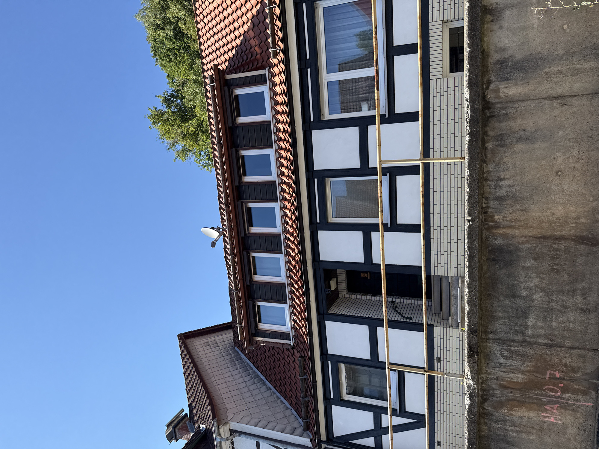 The image shows a close-up, low-angle view of the facade of a traditional half-timbered house, likely located in Bad Lauterberg im Harz, Germany. The house features a dark wooden timber frame with white infill panels. The upper floor has several windows with dark frames, and the roof is covered with red tiles. A satellite dish is visible on the roof. A yellow railing runs horizontally across the front of the house, above a section of white brickwork. Below the house, a weathered concrete wall is visible with pink graffiti that reads "HA | 0.7". The sky above is clear and blue. The environment suggests a residential area, possibly on a slope, given the visible retaining wall.