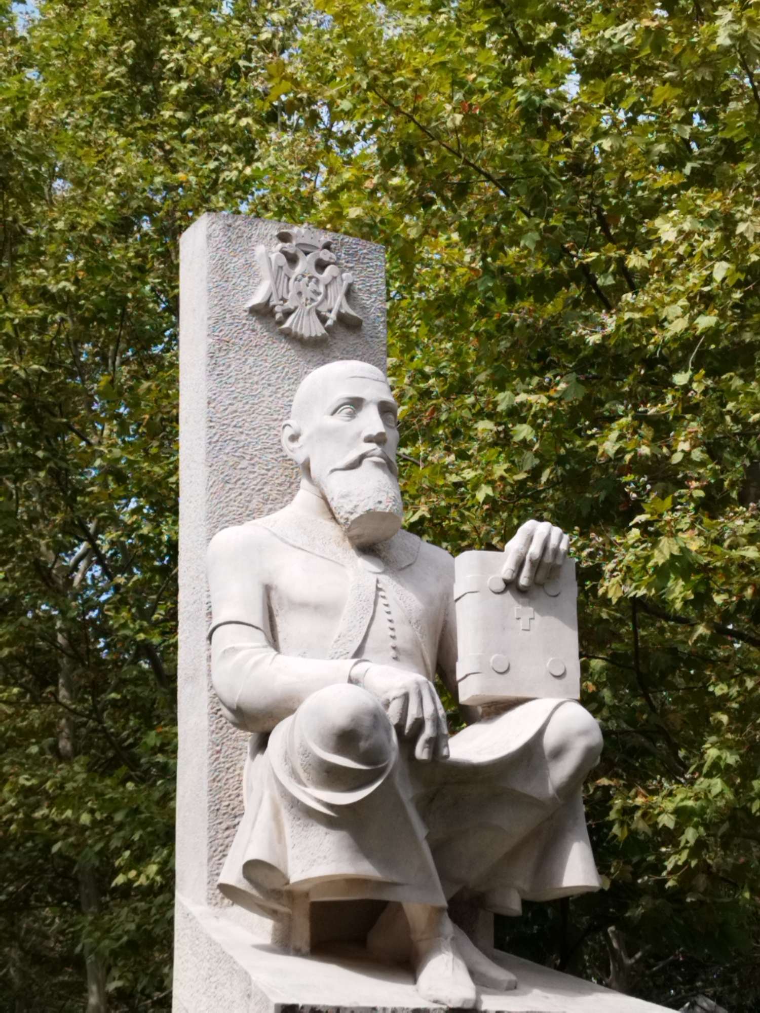 A white stone statue of a seated man is positioned in front of a backdrop of lush green trees. The man has a beard and is wearing what appears to be historical attire. He is holding a book-like object in his right hand. Above the man's head, carved into the stone structure behind him, is an emblem featuring two eagles and a crown, which resembles the coat of arms of Romania. The statue is located in Bucharest, Romania, specifically in the Carol Park, in front of the monument dedicated to Carol I of Romania.