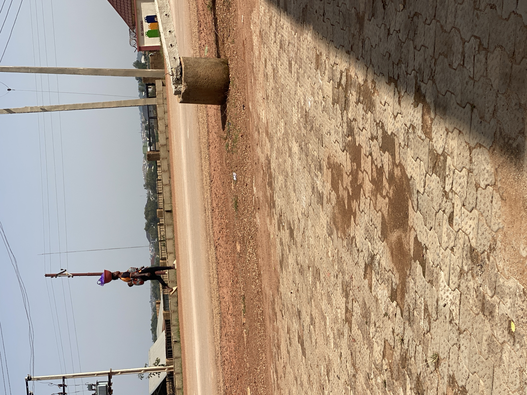 A woman walks along a road in Jos, Nigeria, balancing a large red bowl filled with items on her head. She also carries a baby strapped to her back. The road is a dusty, reddish-brown color, with a paved walkway on the side. Several electrical poles and wires are visible in the background, along with some buildings and trees. The sky is clear and blue, suggesting a sunny day.