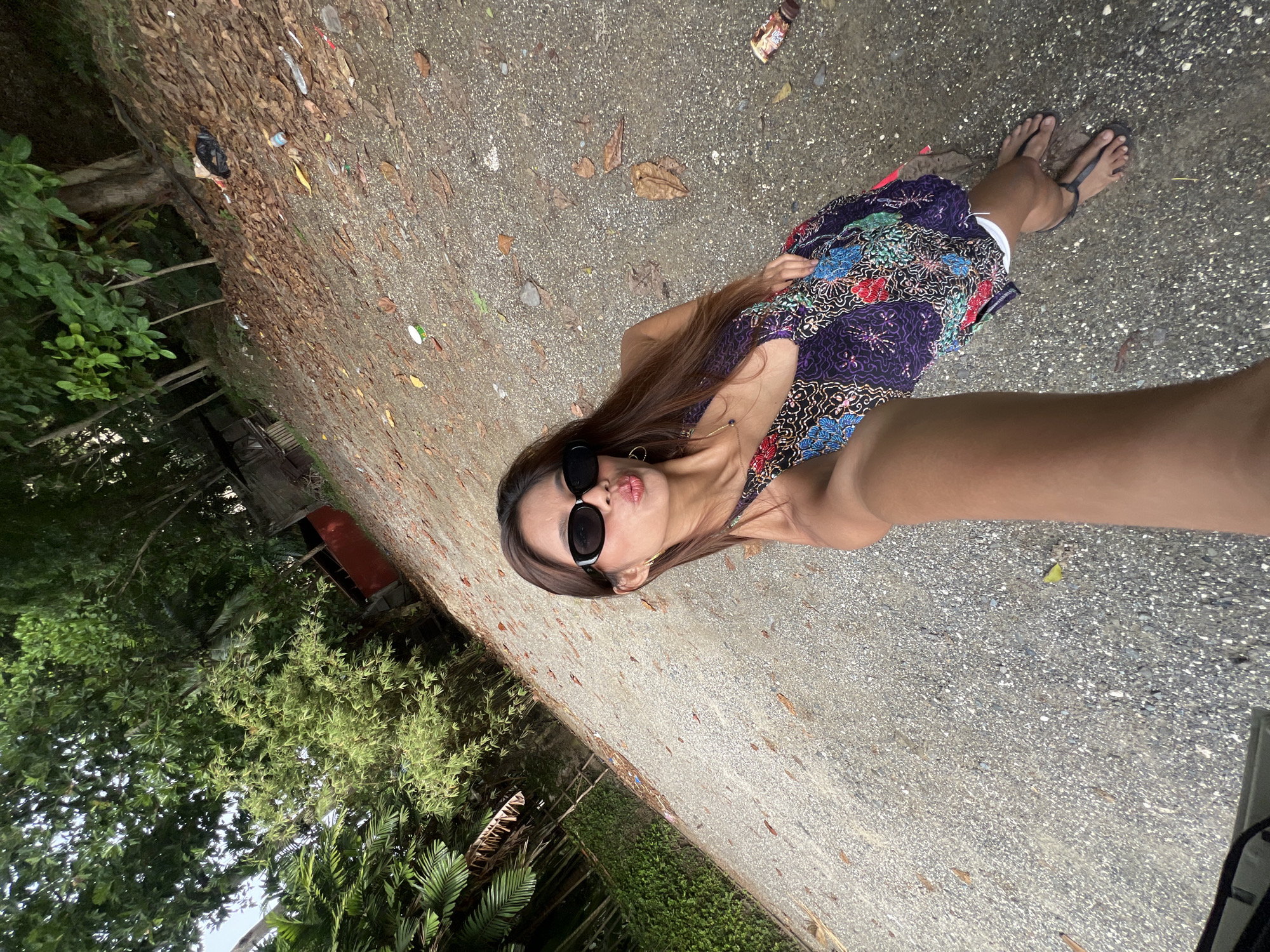 A young woman is posing for a selfie in Banaybanay, Philippines. She is wearing a patterned dress and sunglasses. She is standing on a gray, concrete ground next to a line of trees. Some litter is strewn along the edge of the concrete near the foliage. The background includes a patch of trees, and part of a weathered, red structure. The woman's arm is outstretched, holding the camera.
