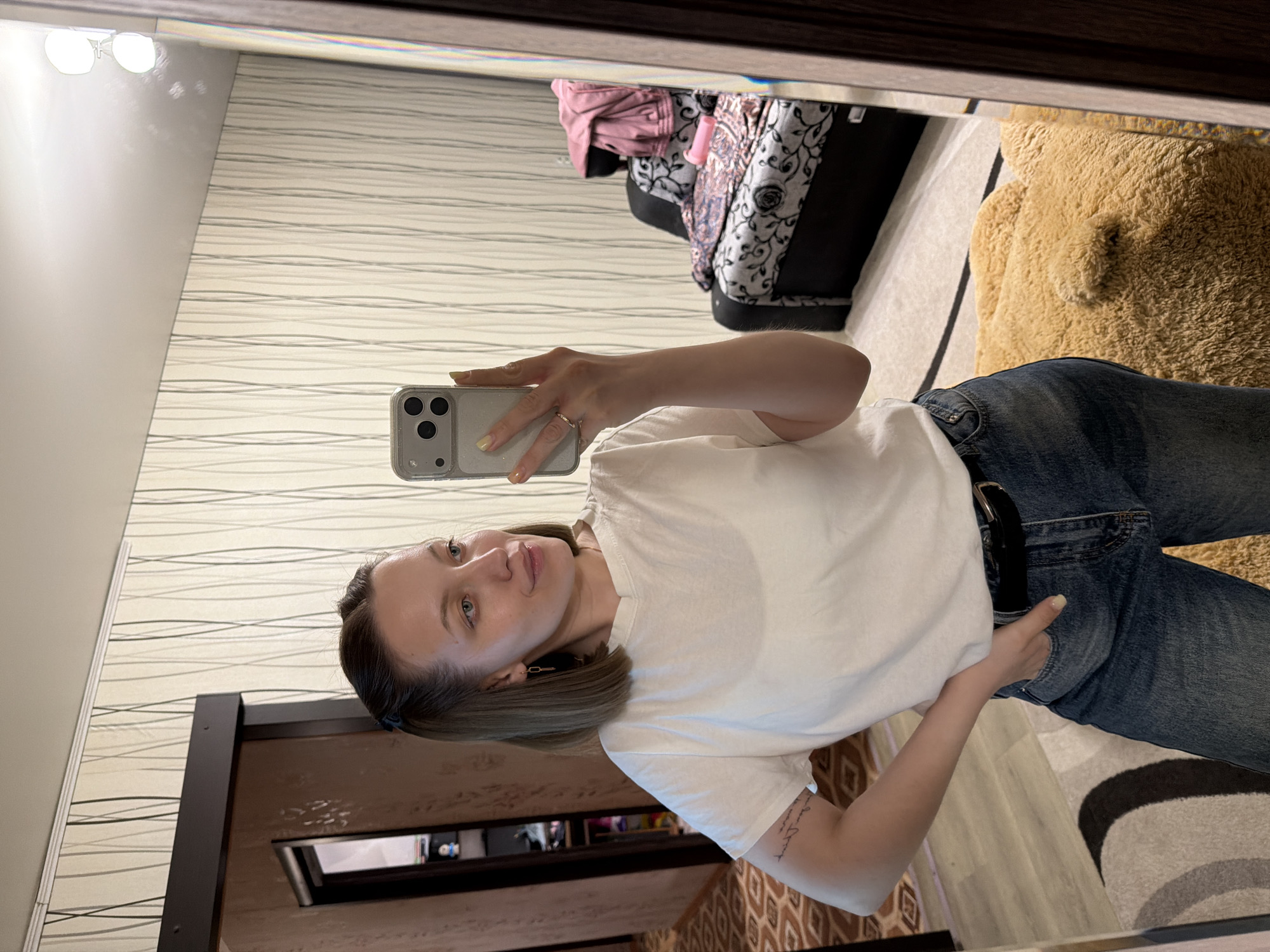 A young woman is captured in a mirror selfie taken indoors, likely within a private residence in Reshetnikovo, Russia. She has fair skin, light-colored eyes (blue/green), and shoulder-length brown hair with lighter, possibly ombre, ends. She is dressed in a plain white short-sleeved t-shirt, blue denim jeans, and a dark belt. Her left hand, adorned with light yellow nail polish and a ring on the ring finger, holds a light-colored smartphone with a triple-camera setup and a clear, slightly glittery case. Her right hand is casually placed in her jeans pocket, revealing a tattoo on her right forearm, the text of which is illegible. Small earrings are visible on her ears.
The background, reflected in the mirror, features a light-colored wallpaper with subtle horizontal wavy line patterns. To the upper right, a bed or couch is visible, covered with various textiles including pink and patterned fabrics. To the lower right, a large, fluffy brown object, resembling a teddy bear or a plush rug, is situated on a patterned area rug. A dark wooden door frame is seen in the lower left portion of the image. Overhead, bright light fixtures are reflected. The woman's expression is neutral to slightly serene as she looks at her reflection. The setting suggests an ordinary moment in a well-lit home environment.