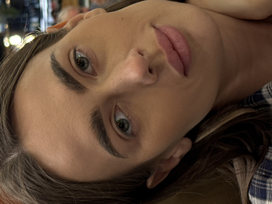 A close-up, slightly off-center portrait captures a young adult female in an indoor setting. She possesses brown hair with lighter highlights and some visible gray strands near the part, dark and well-defined eyebrows, green/hazel eyes gazing slightly upward and to the right, a straight nose, and full, naturally colored lips. A blue and white plaid garment is visible on her lower left shoulder. Her right hand appears positioned near her neck or lower jawline. The background is blurred with a shallow depth of field, revealing out-of-focus artificial lights and indistinct shapes suggestive of shelving or displays, along with a faint, blurred human form (likely a head) on the far right. This scene is located in the city of Verona, Italy.