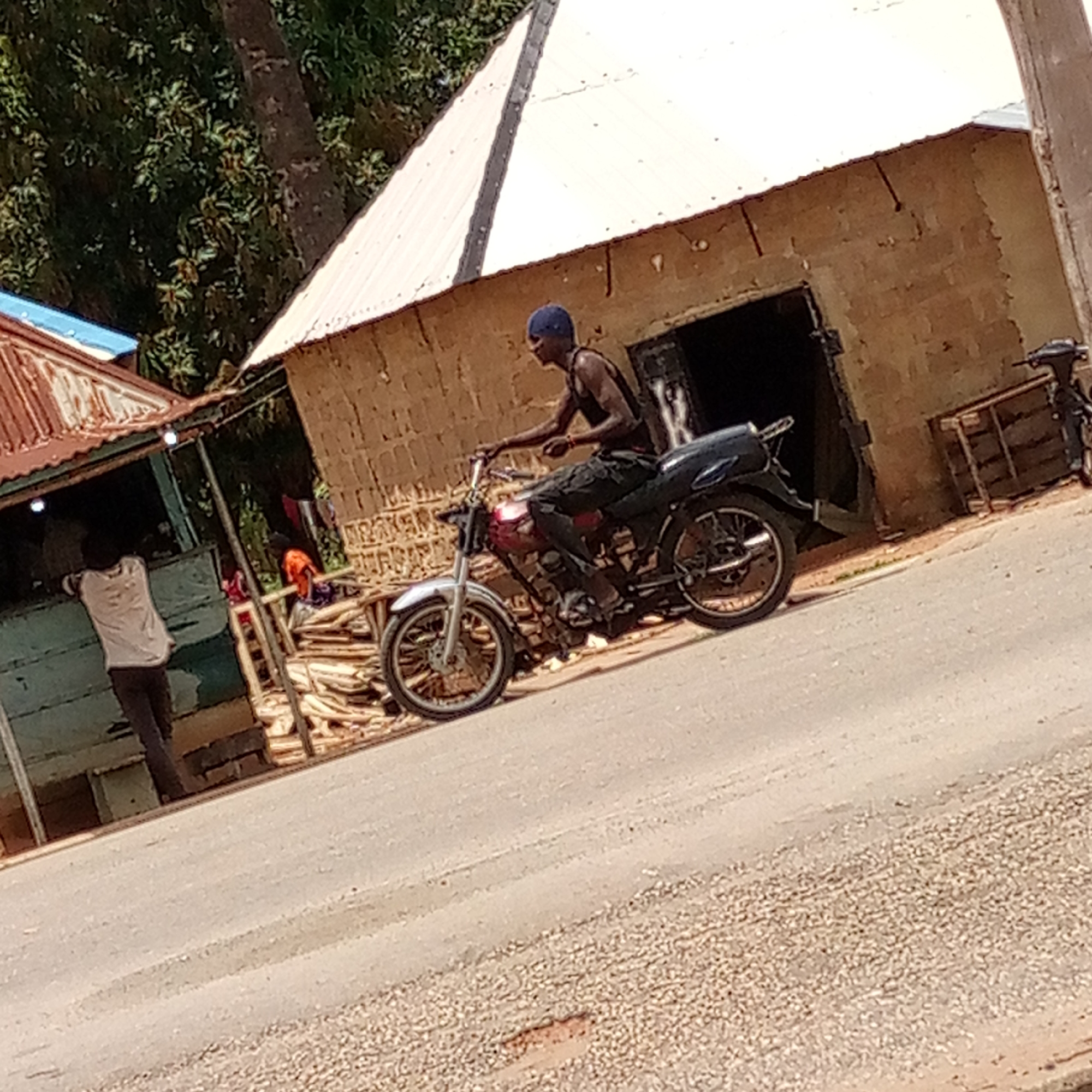 The image depicts a man on a motorcycle on a paved street in Mararaba, Nigeria. The man is wearing a dark tank top and dark pants. He has a blue cap on his head. The motorcycle is red and black. In the background, there is a building with a white, corrugated metal roof. To the left of the building, there is a small shop with a blue-painted exterior. A man in a white shirt and dark pants stands near the shop's entrance. There are stacks of what appear to be wooden planks or boards near the shop. The street is partially paved and appears worn. Another motorcycle is parked in front of a building with an open door.