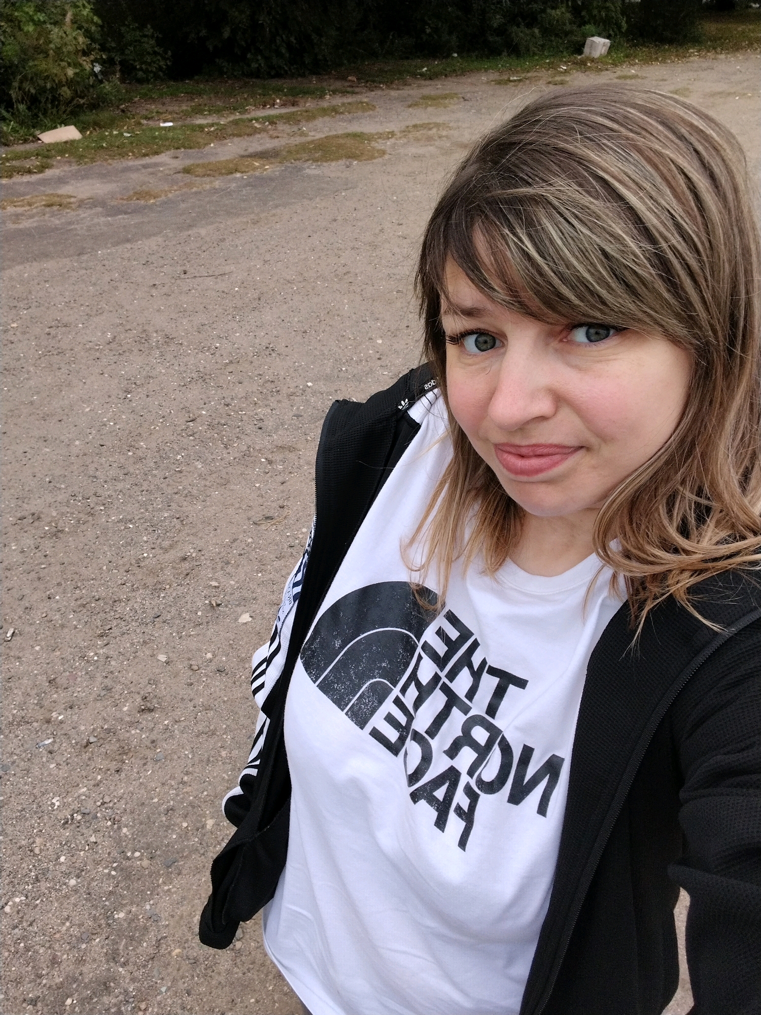 The image is a close-up, high-angle selfie of a woman with brown and blonde hair and blue eyes, wearing a black jacket over a white t-shirt with a large black logo that reads "THE NORTH FACE". She is outdoors, with a dirt and gravel road filling the background. There is some green foliage and trees in the upper background, and what appears to be a discarded piece of cardboard or trash on the left side of the road. The woman is looking at the camera and smiling slightly. The scene appears to be in a rural or semi-urban area. There is no specific indication of the location being Yaroslavl, Russia.