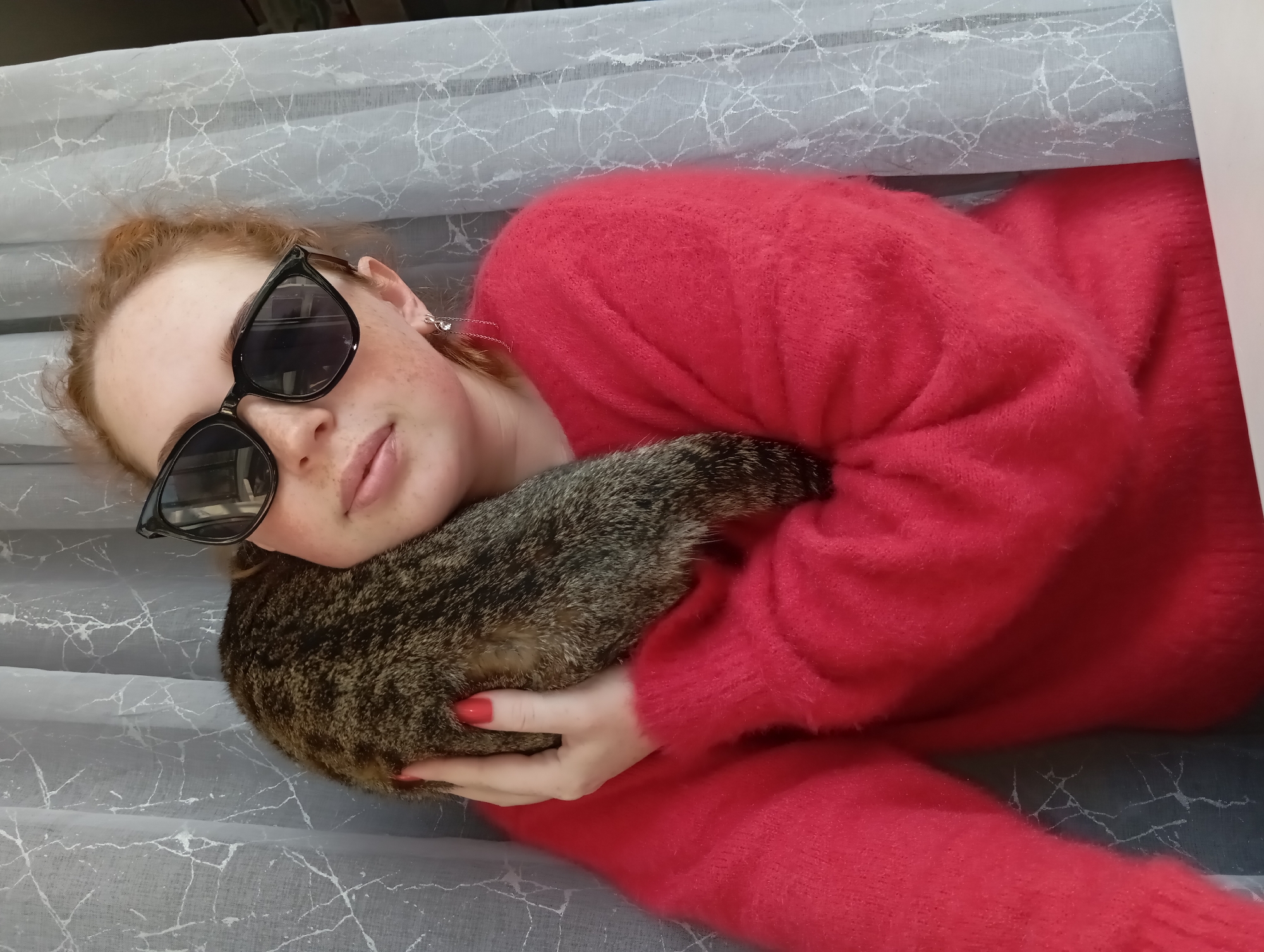 A person with red hair, wearing black sunglasses and a red sweater, is holding a tabby cat. The person is lying on a surface with a light grey marble-like pattern. The scene might be indoors. No specific information confirms the location as Yurgamysh, Russia.