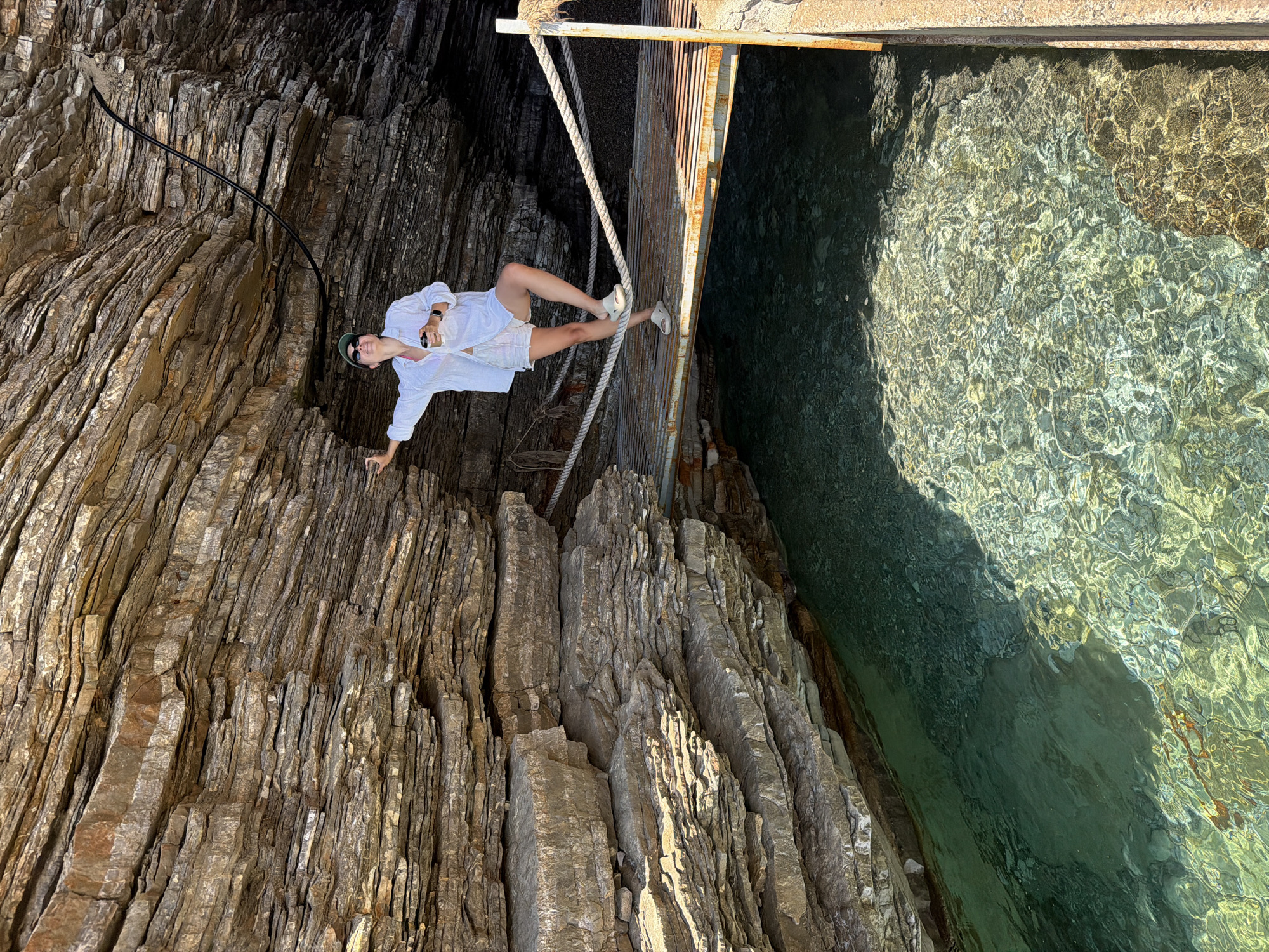 A person is standing on a metal platform suspended between jagged rocks and clear water. The person is wearing a white shirt, white shorts, and white shoes. They have a watch on their wrist and a small object in their hand. A black cable runs along the rock face to the left of the person. Two ropes hang from the platform. The clear water shows light refractions on its surface. The rock formation consists of stacked, sedimentary layers. This location is in the city of Budva, Montenegro.