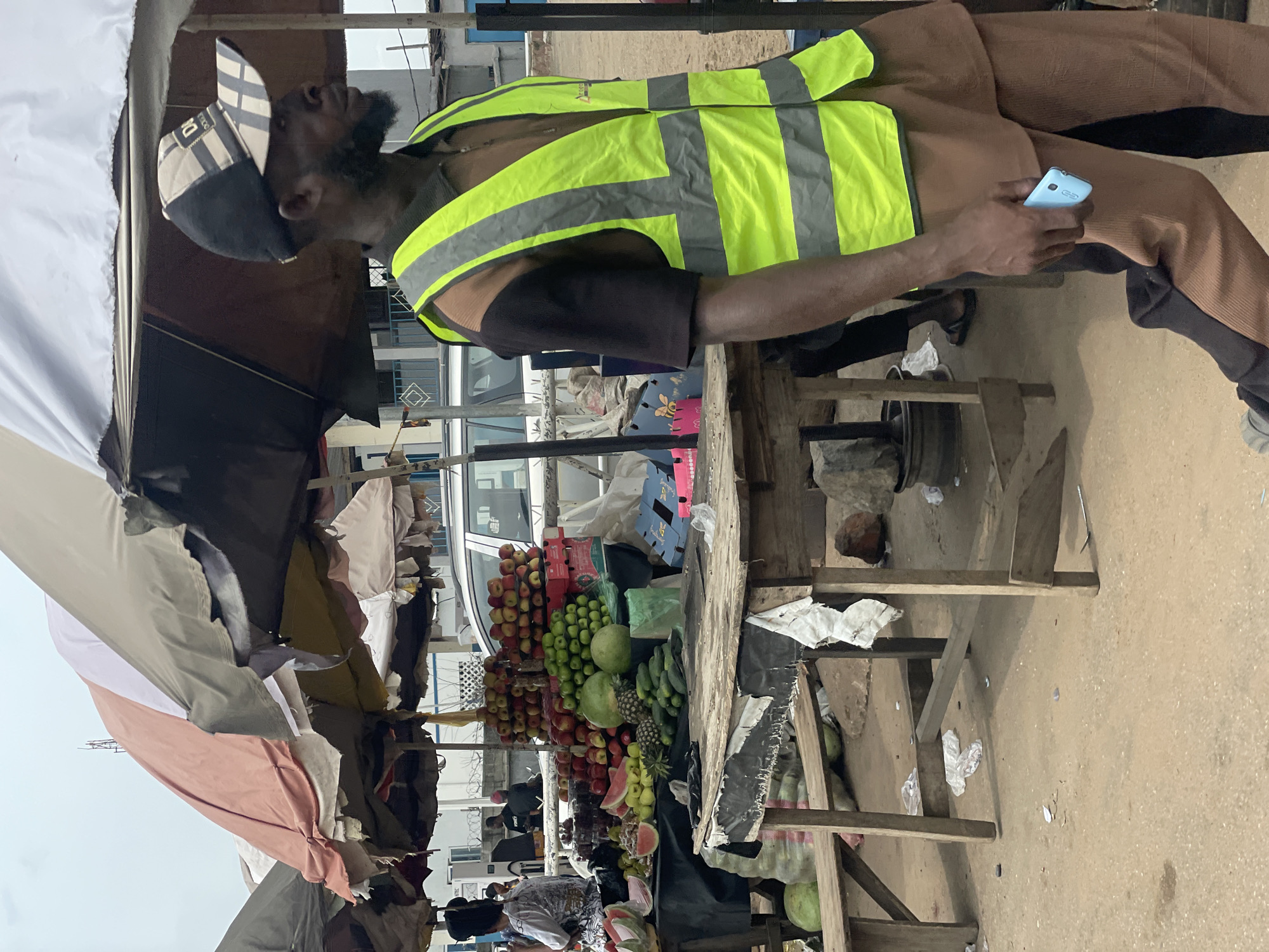 A man wearing a bright yellow reflective vest over a brown shirt and brown pants walks on a dirt road in front of a market stall filled with fruits and vegetables. He is holding a light blue cell phone in his right hand. The market stall is covered by a striped umbrella and has various fruits such as apples, pineapples, watermelons, and cucumbers displayed on wooden tables. In the background, a silver SUV is parked and people are visible behind the market stall. The scene appears to be in Lokoja, Nigeria, characterized by its open-air market setting and dusty environment.