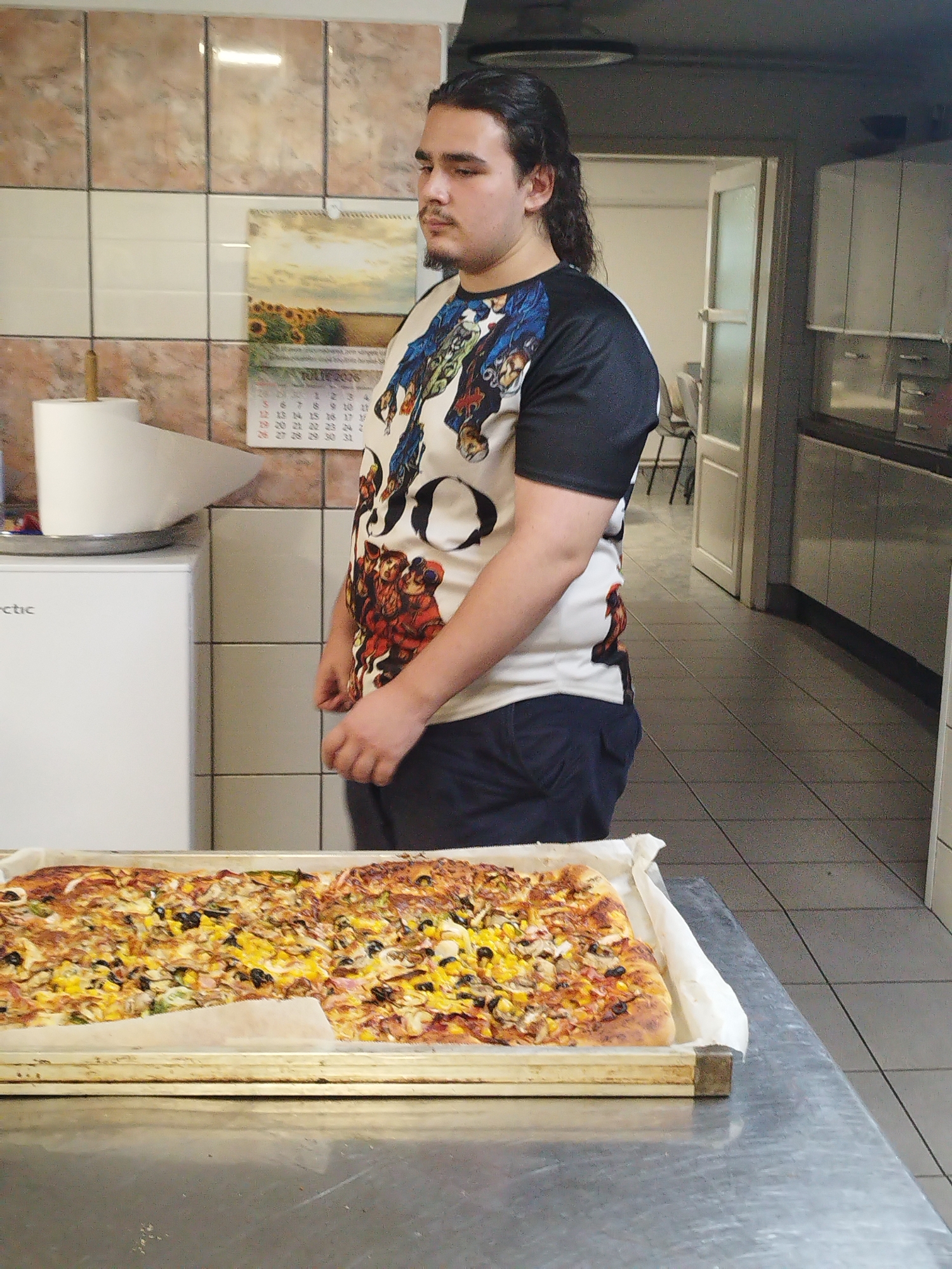 The image depicts a young man with dark, medium-length hair, wearing a t-shirt with a graphic print, standing in what appears to be a commercial kitchen. In the foreground, a large rectangular pizza is presented on a baking tray lined with parchment paper. The pizza is topped with various ingredients including corn, olives, and what appear to be mushrooms and peppers. Behind the man, a roll of paper towels is mounted on a wall. A calendar is also visible on the tiled wall. The environment suggests a food service establishment, likely a pizzeria, located in Bucharest, Romania. The man's posture and gaze indicate he is either posing or being observed.