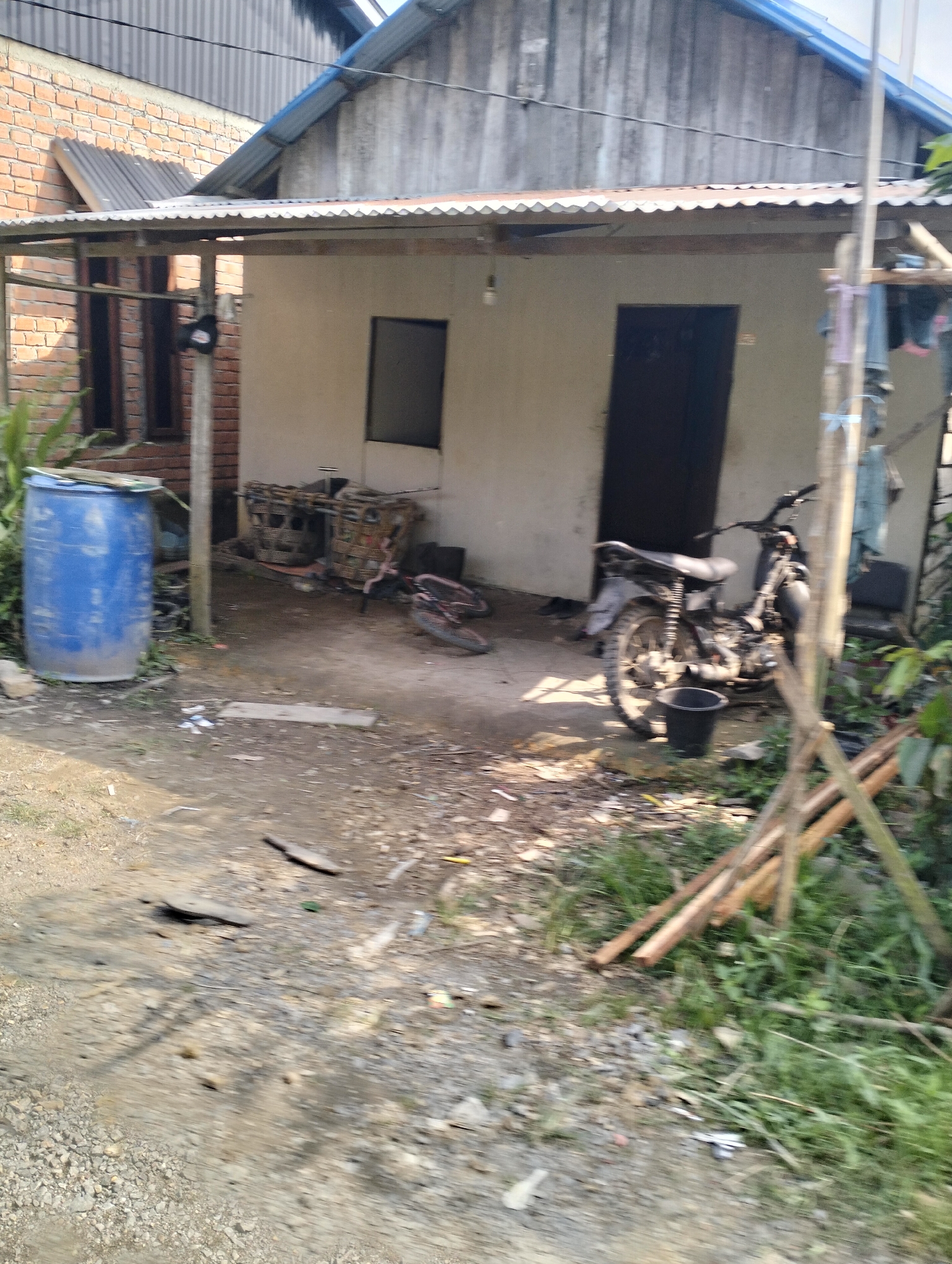 The image depicts an outdoor scene in Serdang Jaya, Indonesia, focusing on a modest house with a corrugated metal roof and light-colored walls. In the foreground, a dirt path leads towards the house, with scattered debris and some vegetation. To the left, a large blue plastic barrel stands near some plants. In front of the house, a black motorcycle is parked. Behind the motorcycle, there are some woven baskets and what appears to be a bicycle frame. The area around the house is unkempt, with visible construction materials like wooden planks stacked on the right side of the image. The overall impression is of a simple, rural dwelling with everyday objects in its vicinity.