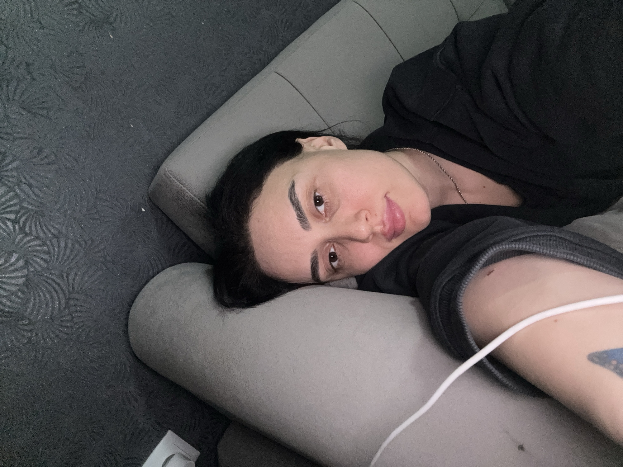 The image features a person, likely female, with dark hair, lying on their side on a light grey cushion or sofa armrest. The person's head rests on the cushion, and they look directly towards the camera with a neutral to slightly contemplative expression. They are wearing a dark-colored top, possibly a hoodie, and a thin silver chain necklace is visible around their neck. A white cable, appearing to be for headphones or charging, runs across their right arm, where a tattoo with a dark blue, wing-like design is partially visible.
The background to the left of the person consists of a dark grey surface with a subtle, repeating pattern resembling swirling foliage or fern fronds, suggesting it could be a textured wall, carpet, or decorative panel. In the bottom left corner, a small white object, possibly an electrical outlet or part of a charging device, is discernible against this patterned surface. The lighting is soft and even, indicating an indoor setting. The scene conveys a casual moment of rest or introspection. The location context is Tbilisi, Georgia.