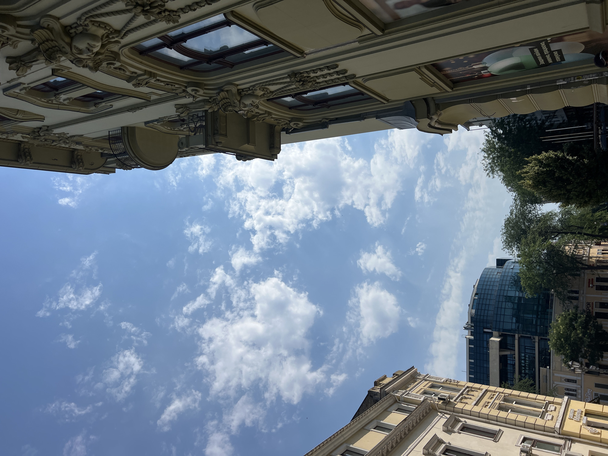 In Odesa, Ukraine, this photograph captures a street scene with a distinct architectural contrast under a bright blue sky dotted with white clouds. The foreground features the ornate facade of a building, likely from the early 20th century, characterized by its creamy yellow hue, detailed plasterwork, including sculpted faces and decorative garlands, and arched windows. To the left, another building with a similar yellow facade and classical architectural elements is visible. In the middle ground, a modern, curved building with a glass facade stands out, bearing signs with text including "CONVERSE" and "Dr. Martens". Lush green trees line the street, partially obscuring other structures. The perspective is tilted upwards, emphasizing the sky and the upper portions of the buildings.
