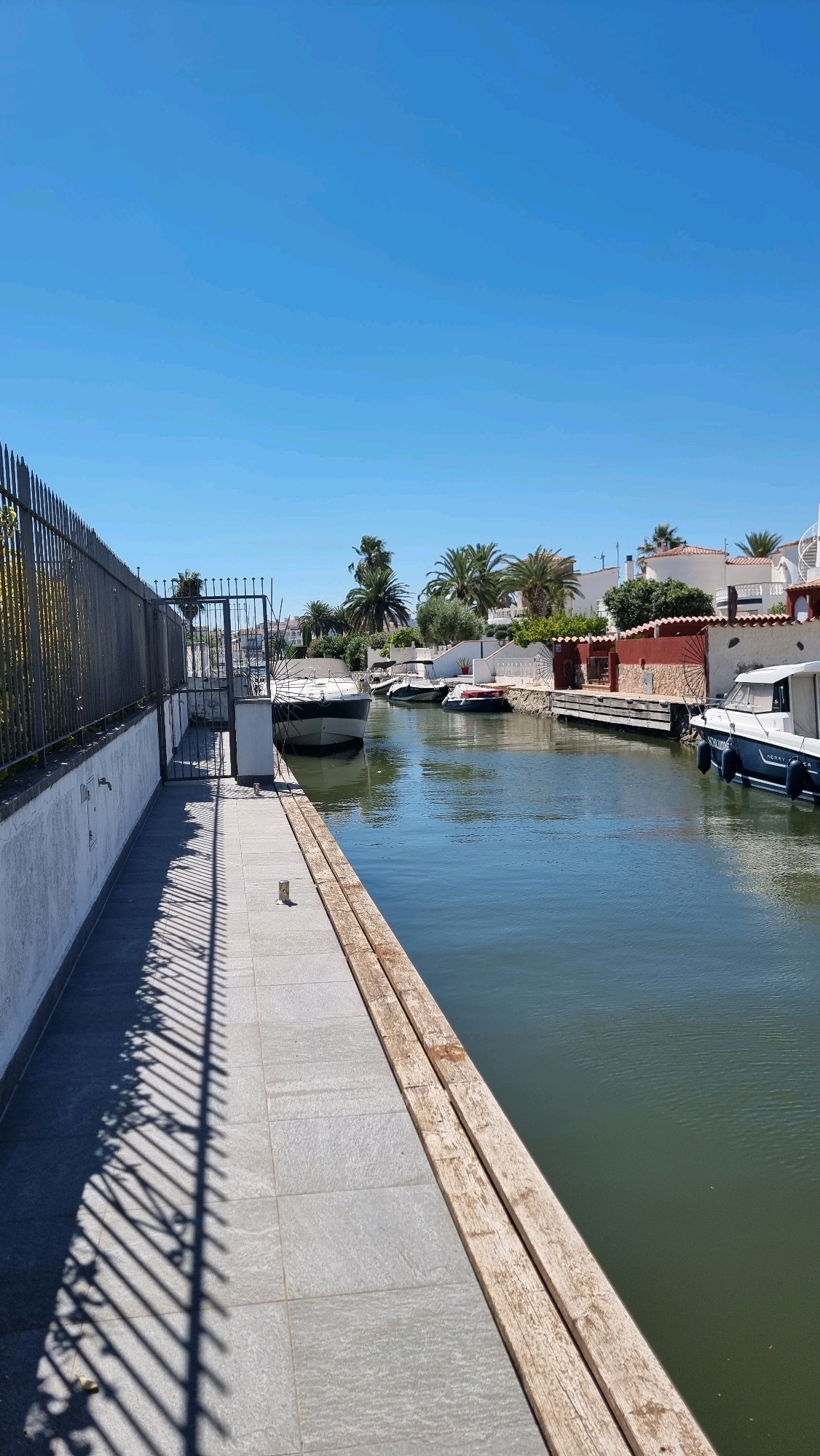The image depicts a canal in Castelló d'Empúries, Spain, on a clear sunny day. The canal runs through a residential area lined with houses and palm trees. Several boats are moored along the canal, including a large white and black yacht, a smaller white boat, and another white boat with a blue trim. The canal is bordered by a paved walkway on the left, separated from the water by a wooden border. A metal fence runs along the side of the walkway, casting a striped shadow. The water in the canal is a dark green color, and the sky is a vibrant blue. The houses have white walls and red-tiled roofs.
