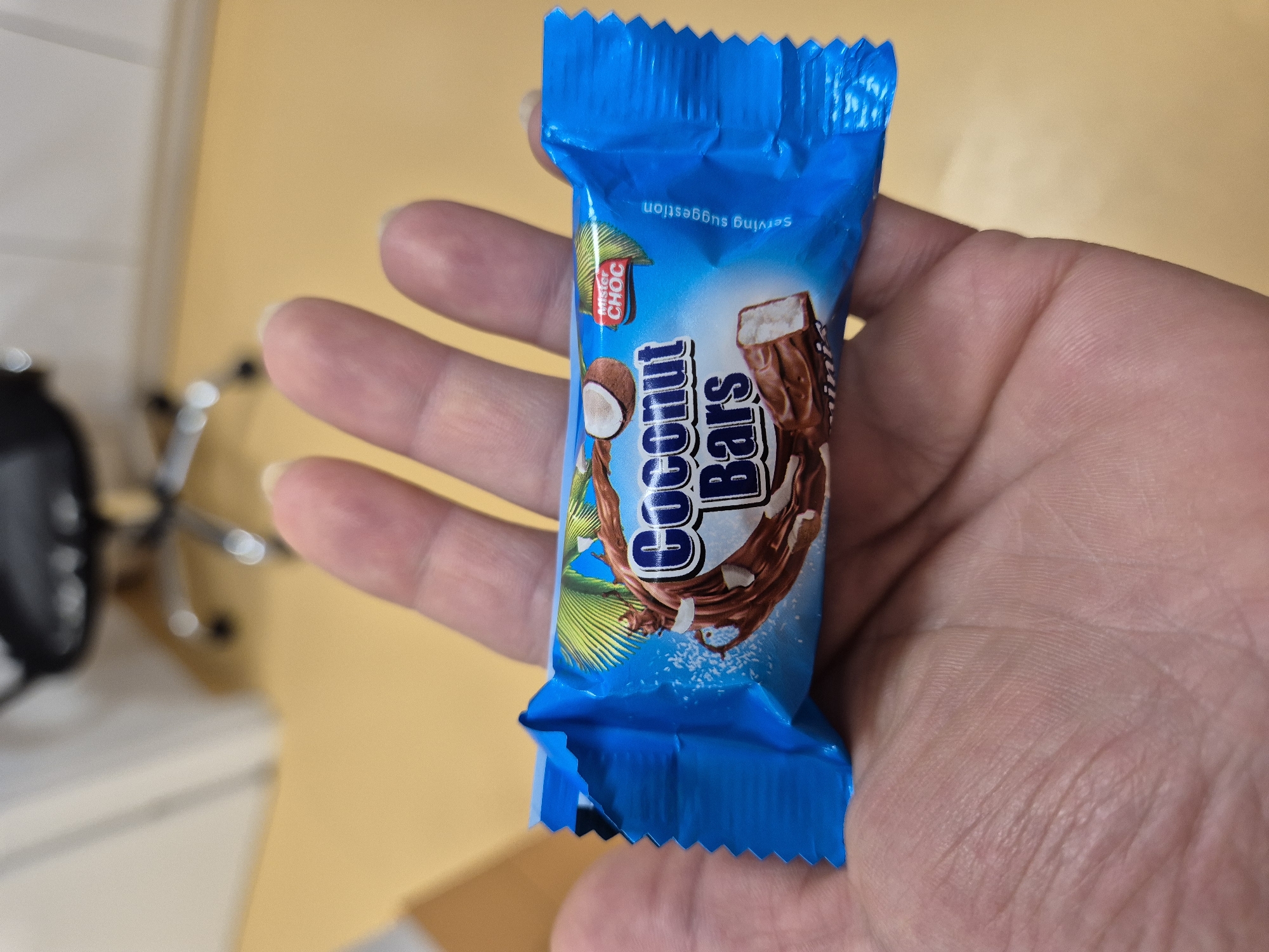 A hand is holding a chocolate bar with a blue wrapper. The wrapper has an image of a coconut and chocolate bar, with the text "Coconut Bars" and "Mister CHOC". The background is blurry and appears to be an office or indoor setting with a yellow floor.