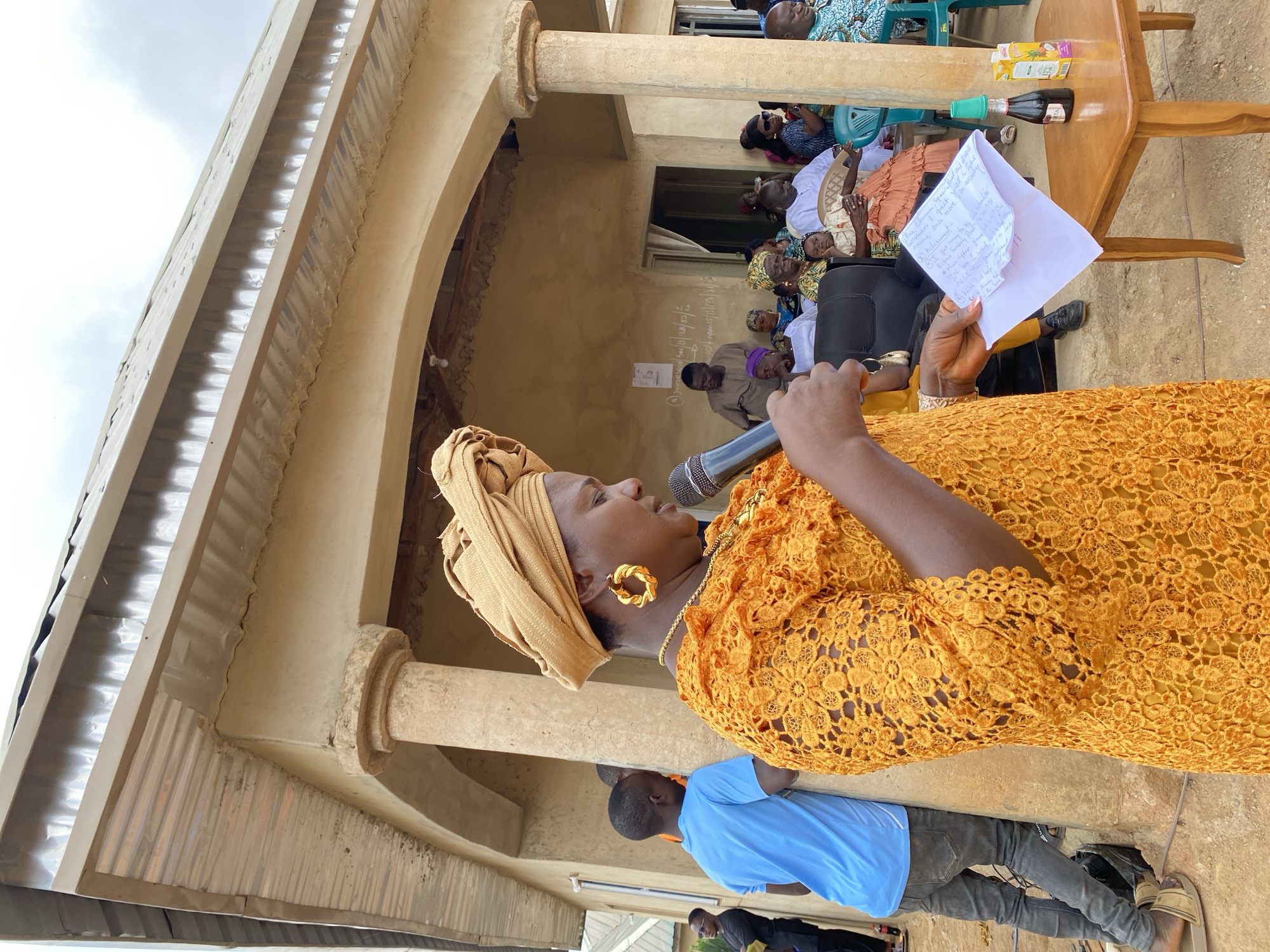 The image depicts a woman in a mustard-colored lace dress and matching headwrap speaking into a microphone while holding a paper in her left hand. She is wearing gold earrings and a necklace. Behind her, seated in chairs, is a group of people. A table with beverages and other items is visible. The event is taking place outdoors, in what appears to be a courtyard or covered area with pillars and an arched entrance. The location is Barikin Sabon Gidan Kanat, Nigeria. A man in a blue shirt stands on the ground in front of her.