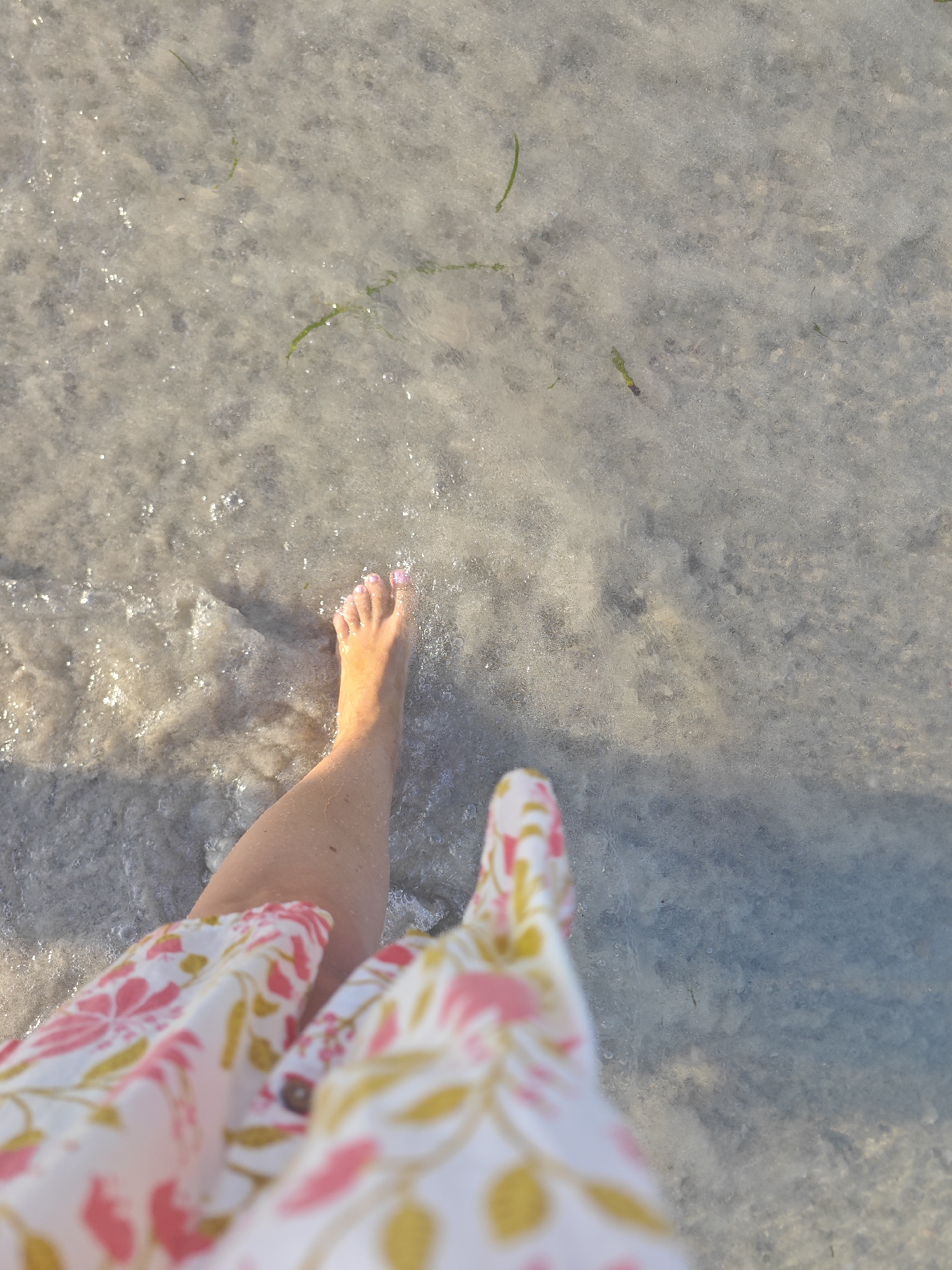 This is a top-down, close-up shot of a person's bare foot and lower leg partially submerged in shallow, clear water. The water appears to be over a sandy or pebbly seabed with some sparse green aquatic vegetation visible. The person's foot is positioned in the lower right quadrant of the image, with the toes and the arch clearly visible. The skin on the leg and foot is lightly tanned, and the toenails are painted with a light pink polish. The person is wearing a garment with a floral pattern in pink and yellow on a white background, which is visible in the bottom part of the frame, obscuring the rest of the body. The lighting suggests it is daytime, with sunlight reflecting off the water's surface, creating small glints and highlights. The environment appears to be a natural body of water, possibly the sea or a shallow lake, consistent with coastal areas like those found near Hiboun, Tunisia.