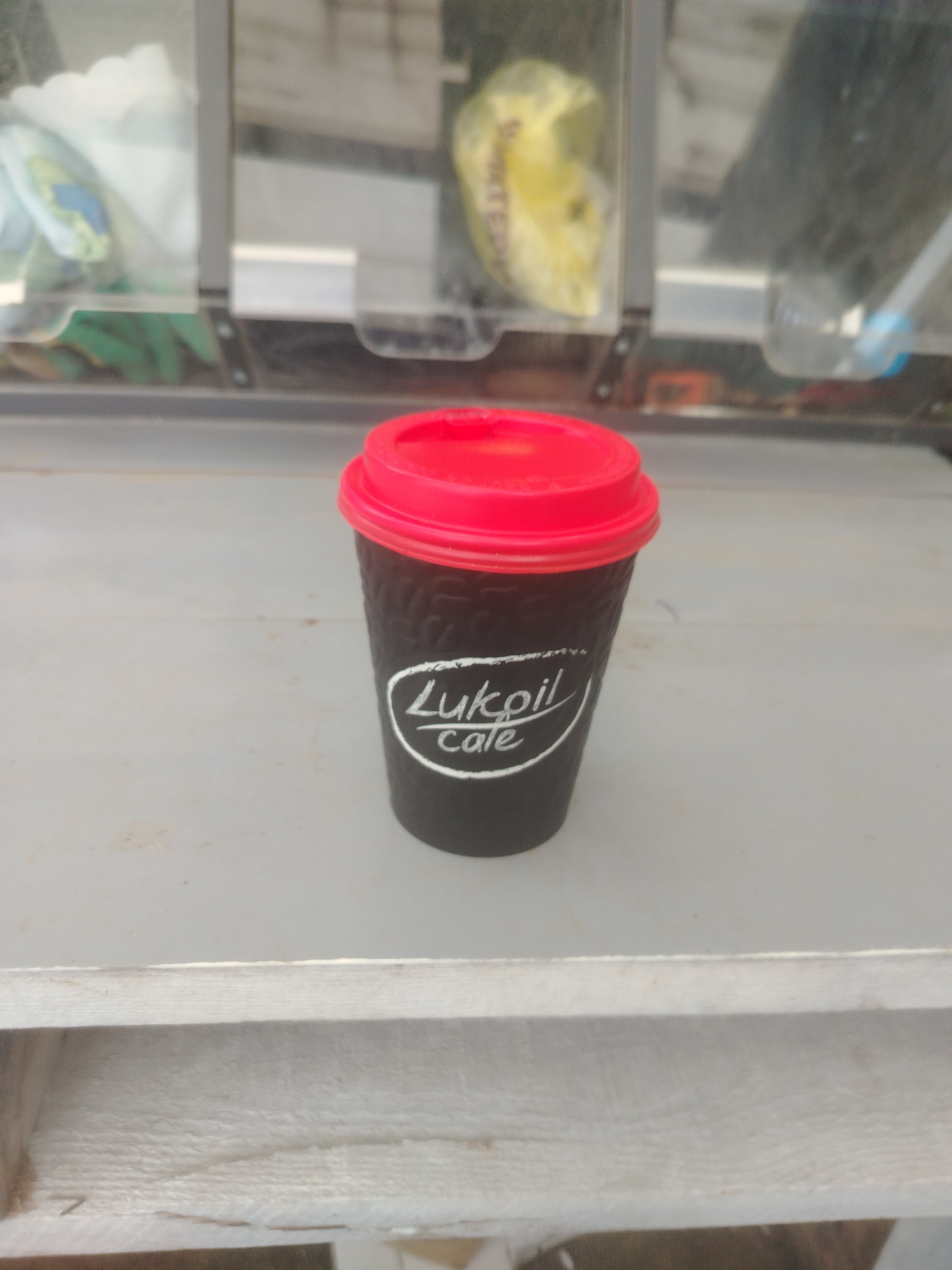 A black disposable coffee cup with a red lid sits on a light-colored surface. White text on the cup reads "Lukoil Cafe". The cup is positioned slightly to the right of the center of the frame. In the background, out of focus, are shelves with various items, including a yellow object in a plastic bag and what appear to be other cups or containers. The environment suggests an indoor setting, possibly a kiosk or a shop. The exact location within Тулаўскі сельскі Савет, Belarus is not discernible from the image itself.