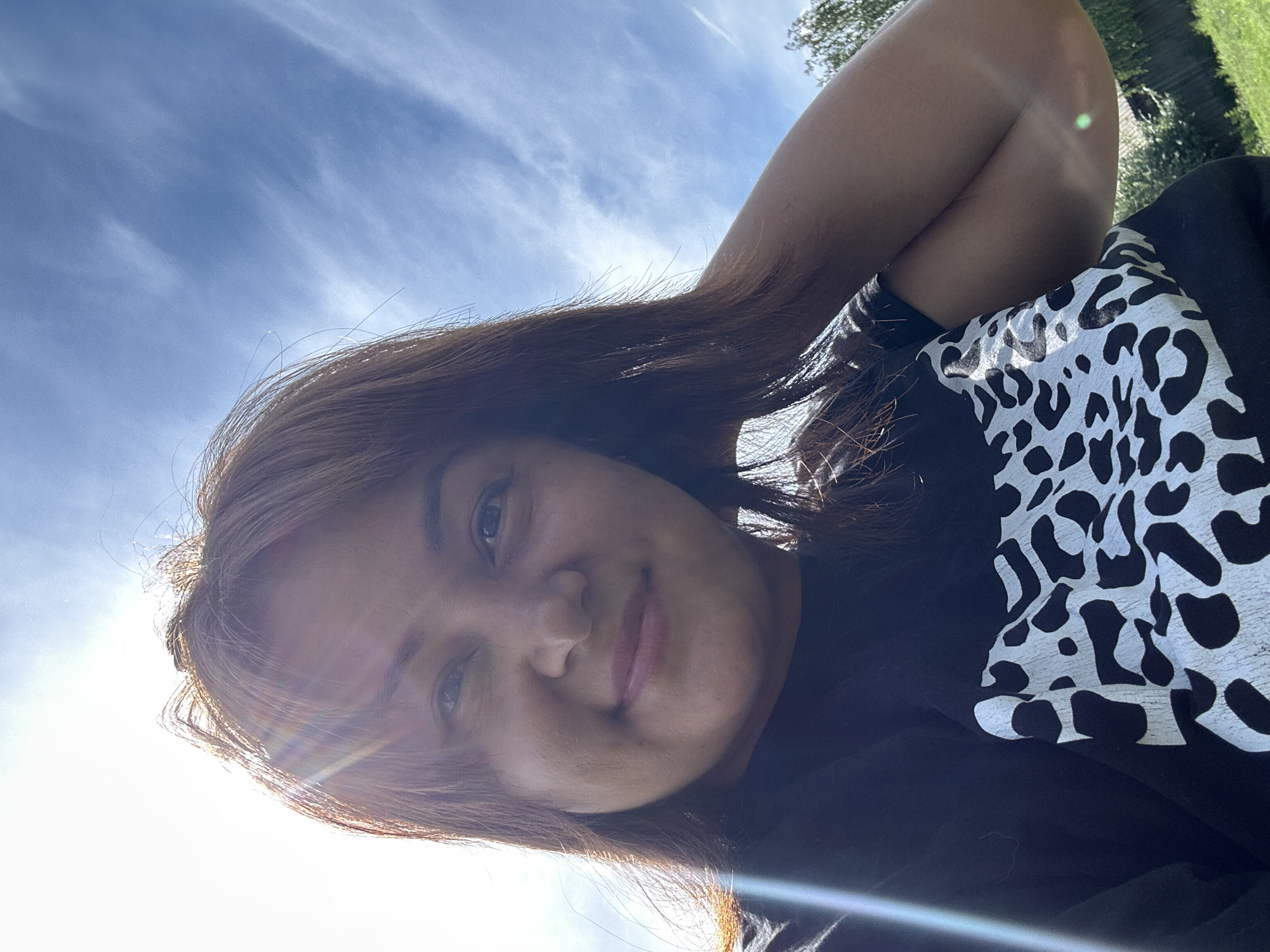 A young woman with medium-length brown hair and brown eyes poses for a selfie outdoors. She is wearing a black t-shirt with a white and black cow print design. The sun is shining brightly, creating a lens flare across the upper left portion of the image. The background consists of a blue sky with thin white clouds and some greenery, including trees and a fence, indicating an outdoor, possibly backyard setting. The woman is looking at the camera with a slight smile, and her right arm is bent and resting behind her head. The image appears to be taken during daylight hours. The location is described as Unknown, Unknown.